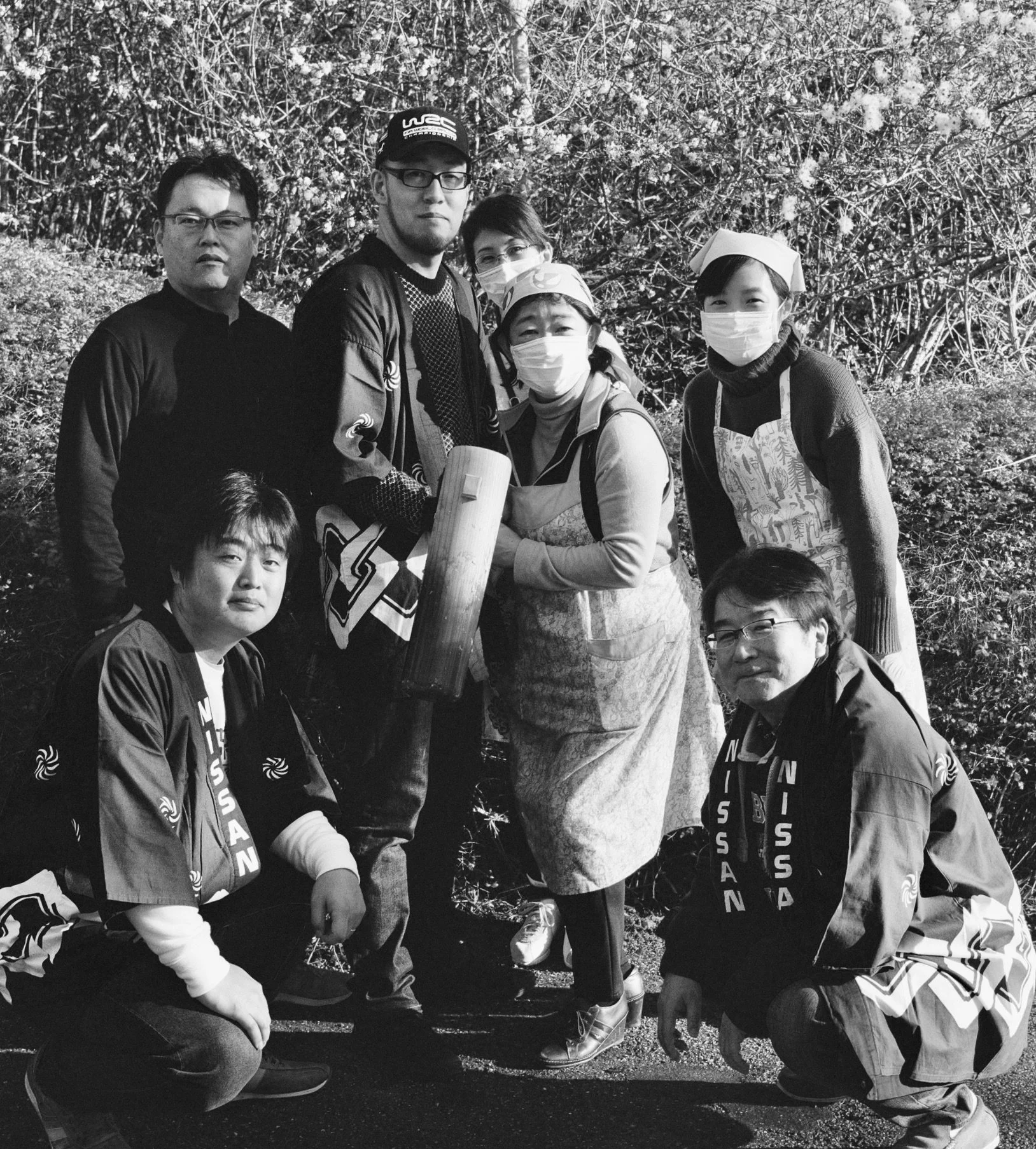 Group of six people outdoors, wearing masks, with three people kneeling and three standing, against a background of dense foliage.