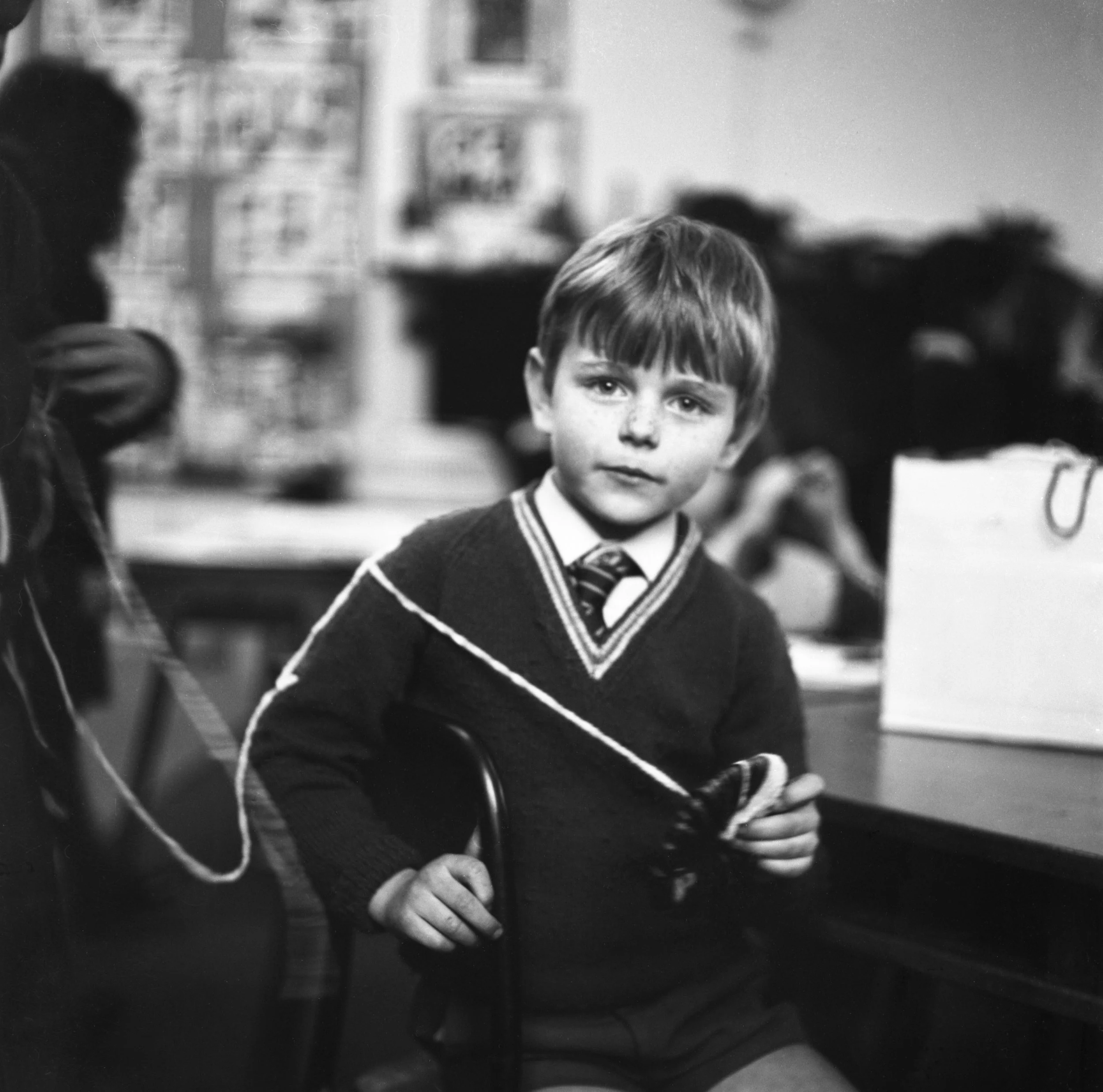 Black and white photo of a young boy with short hair and a serious expression, wearing a sweater, in a classroom setting with other children in the background.