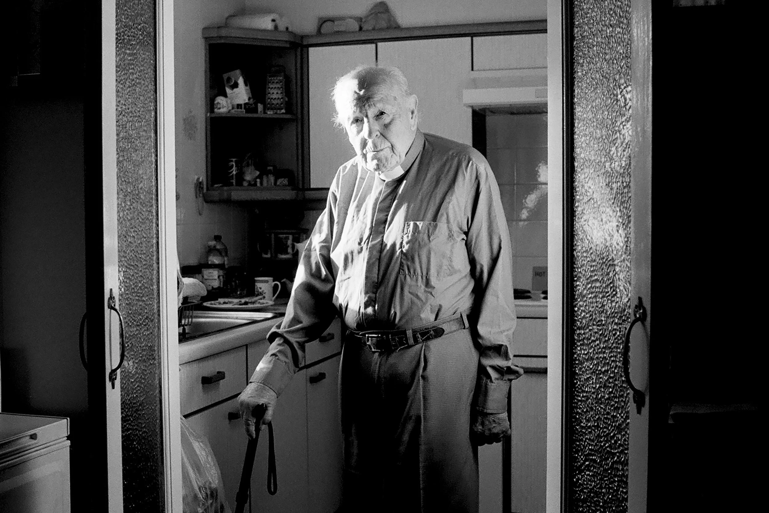 An elderly man with gray hair and a serious expression stands in a kitchen doorway. He wears a light-colored shirt and belt, with one leg propped on a chair. The kitchen has cabinets, a sink, and various food items on the counter, with some shelves a