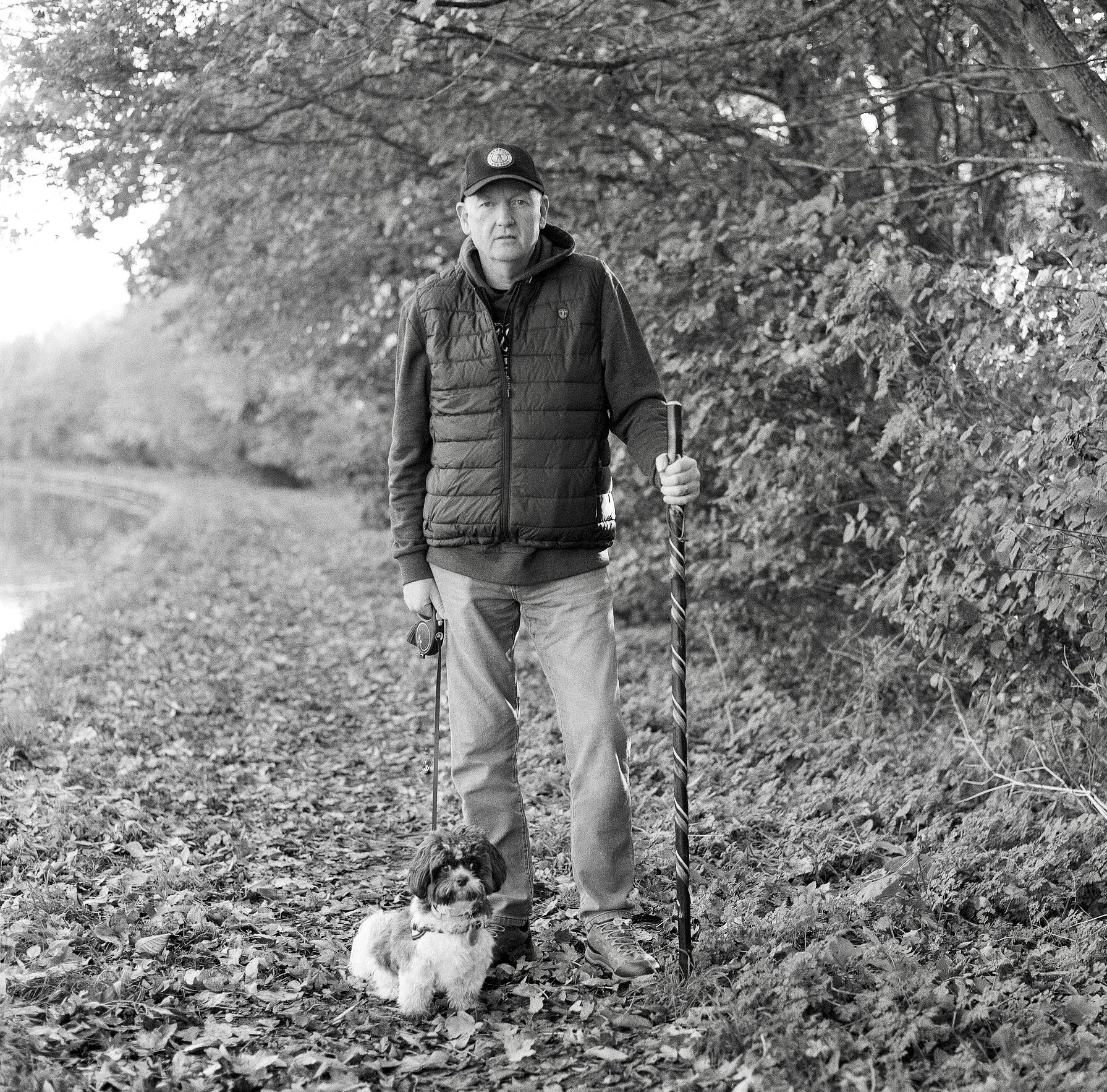 A man walks a dog with a walking stick along a leaf-covered trail in a wooded area, captured in black and white.