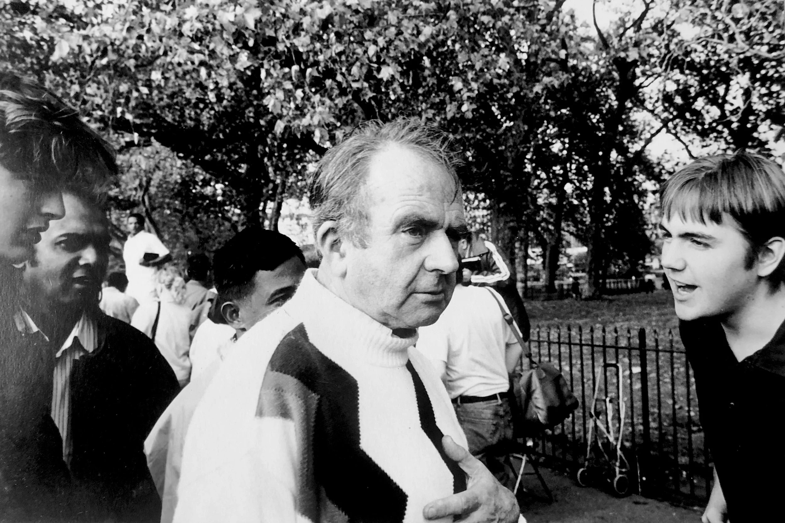 Black and white photo of an older man with gray hair and glasses, speaking to a young man with short hair, outdoors in a park with trees and a fence in the background. Several other people are in the background.