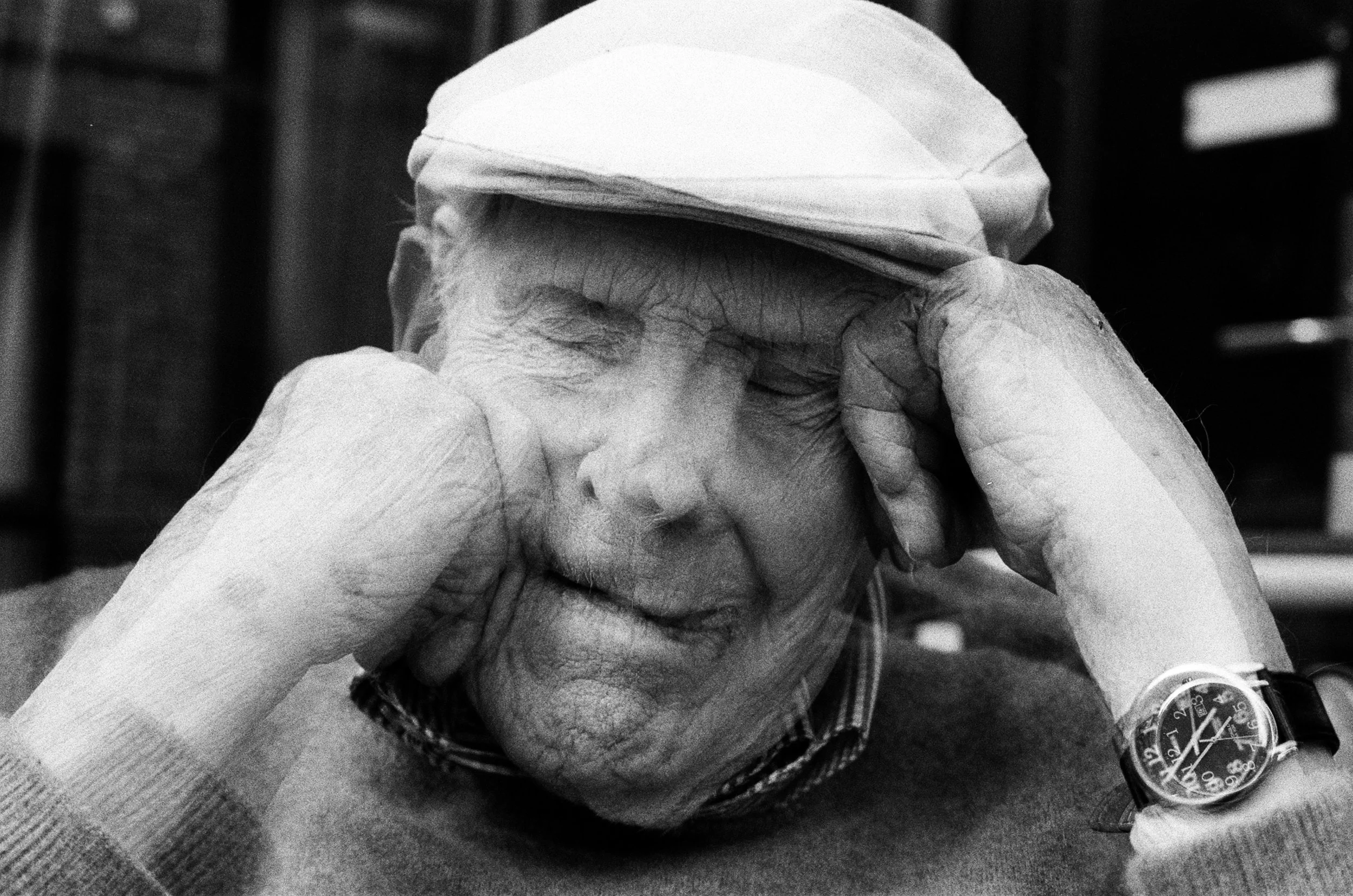 Close-up black and white photo of elderly person with glasses resting their forehead on their hand, which is on a table, wearing a watch, with another hand holding their head. The person appears to be deep in thought or experiencing emotional distres