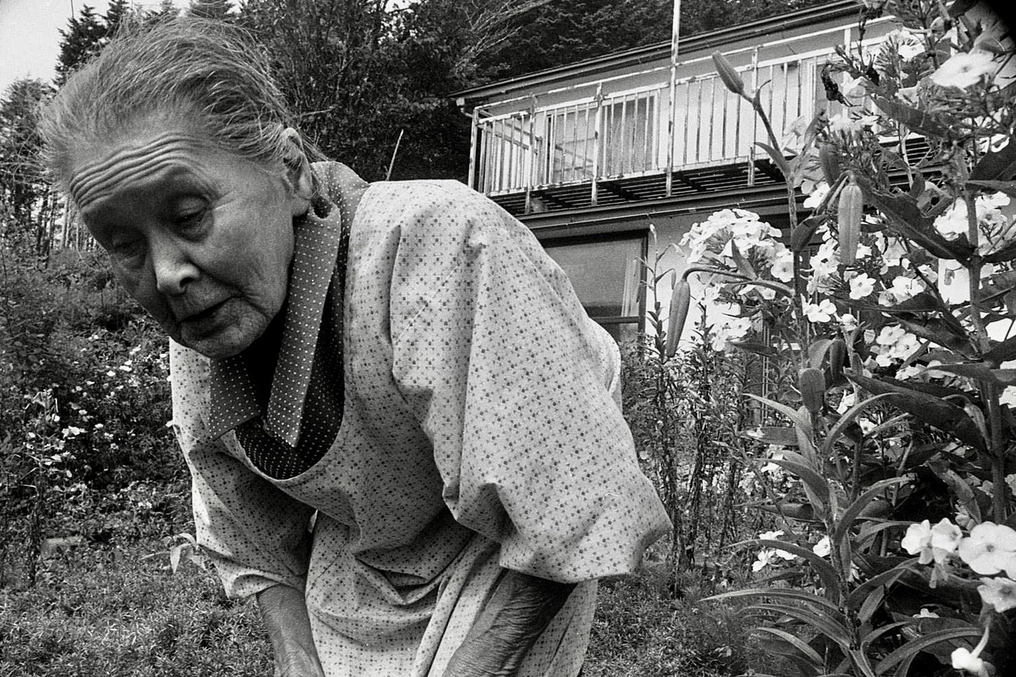 An elderly woman with light-colored hair styled back, wearing a patterned dress with a polka dot scarf around her neck, bends over in a garden with blooming flowers and a house with a balcony in the background.