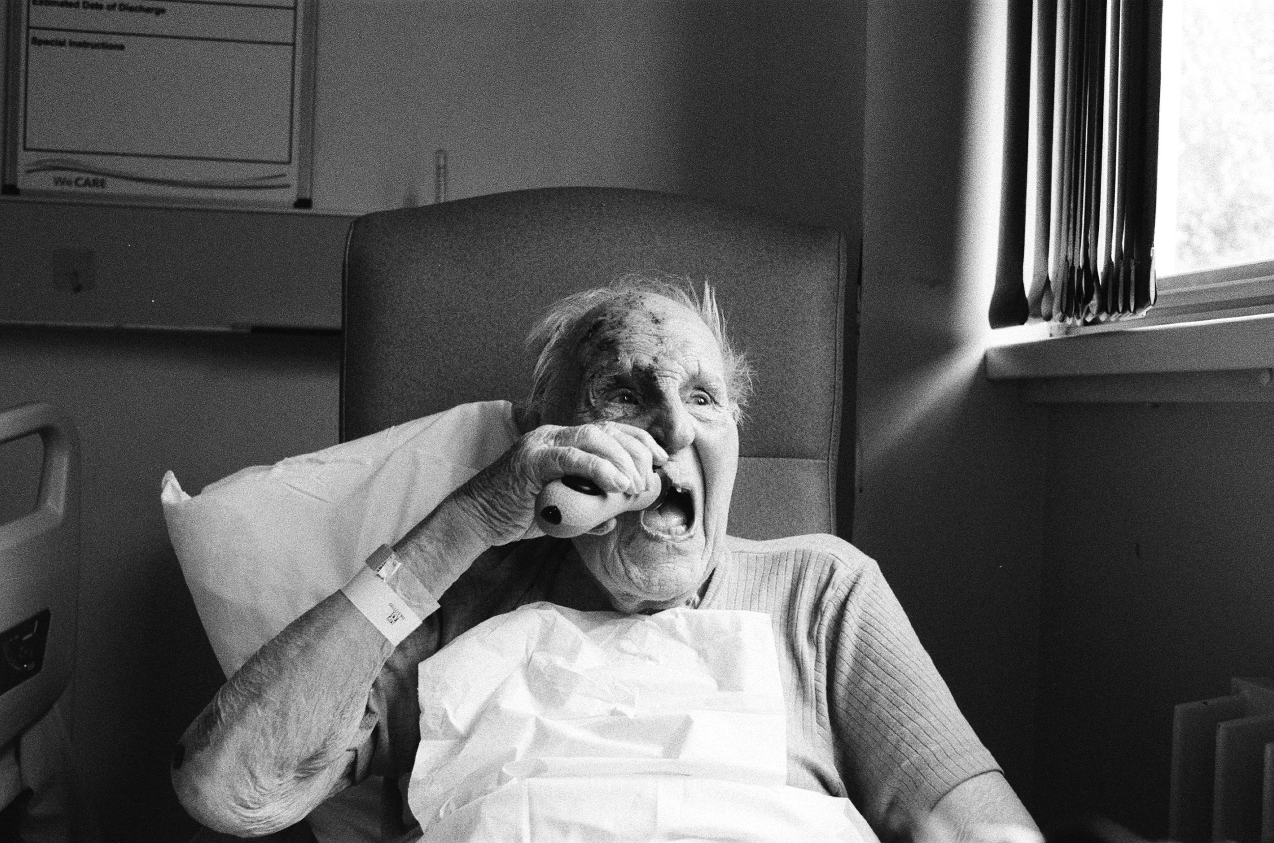 A black and white photo of an elderly woman lying in a hospital bed, looking joyful and holding a small object to her mouth. She has a hospital wristband on her wrist, and there is a hospital room with a window and blinds in the background.