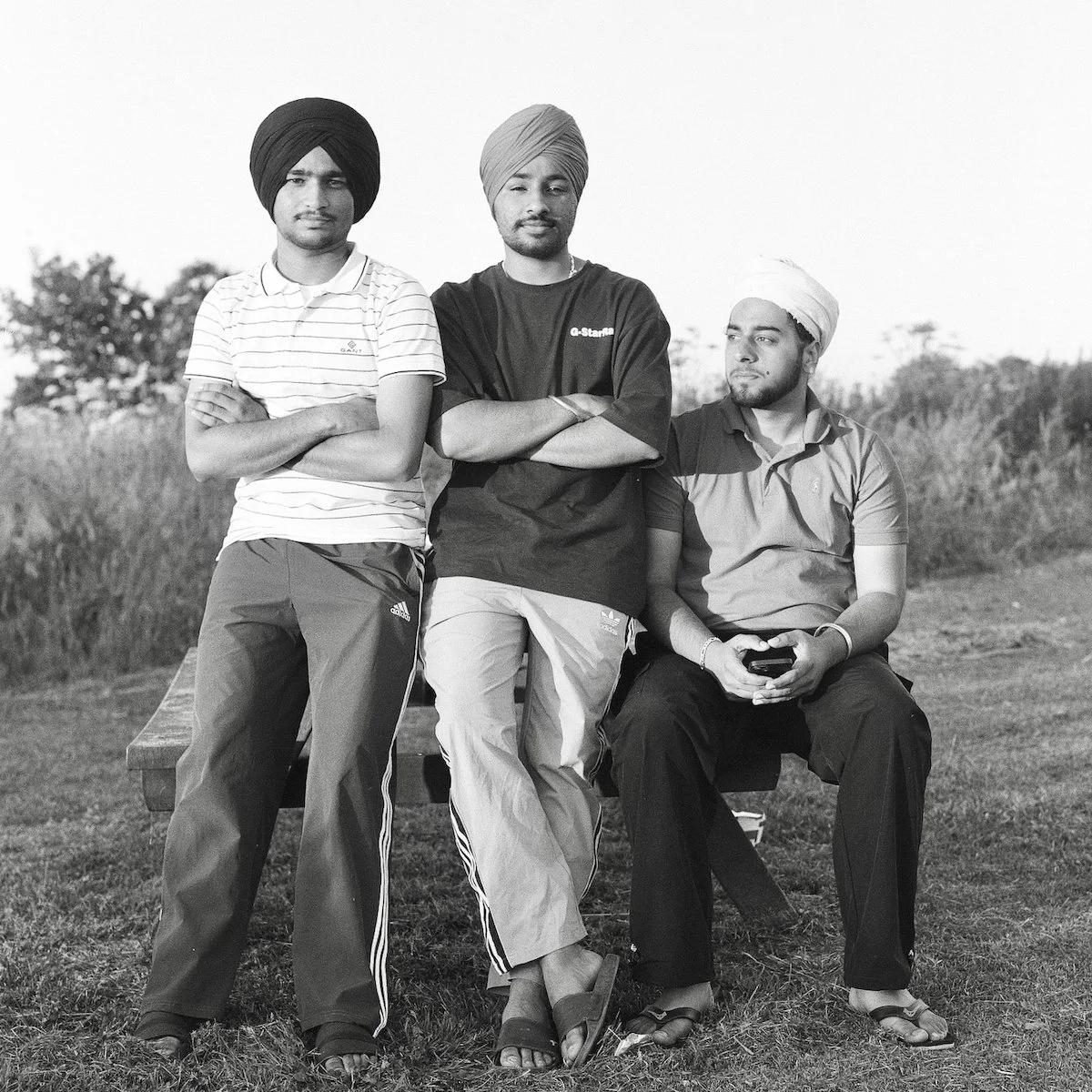 Three men wearing traditional turbans, sitting and standing outdoors on a bench in a field.
