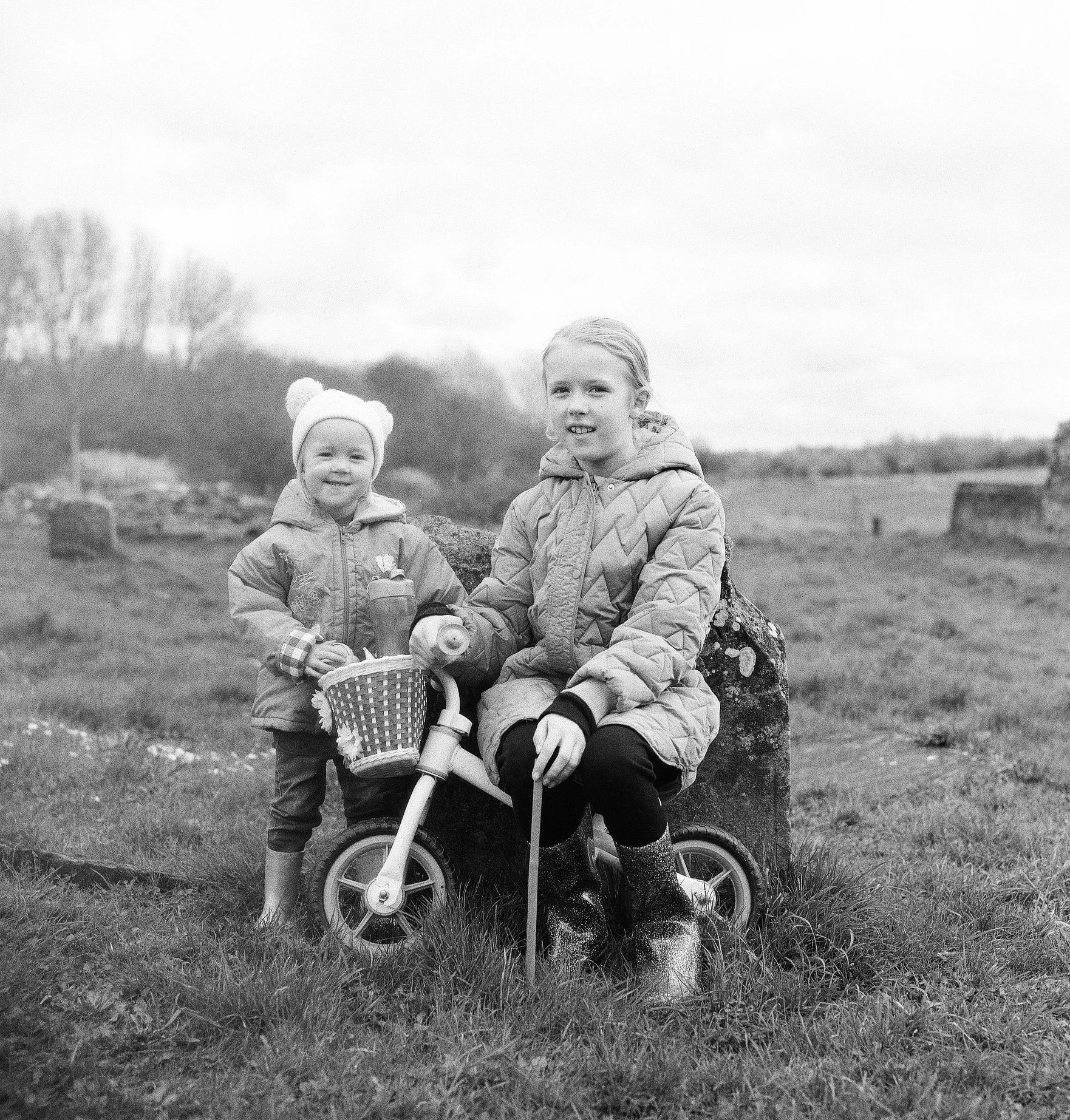 Two young girls outdoors, one sitting on a bicycle and the other standing beside it, both smiling and dressed warmly in jackets, in a rural setting with a grassy field and trees in the background.