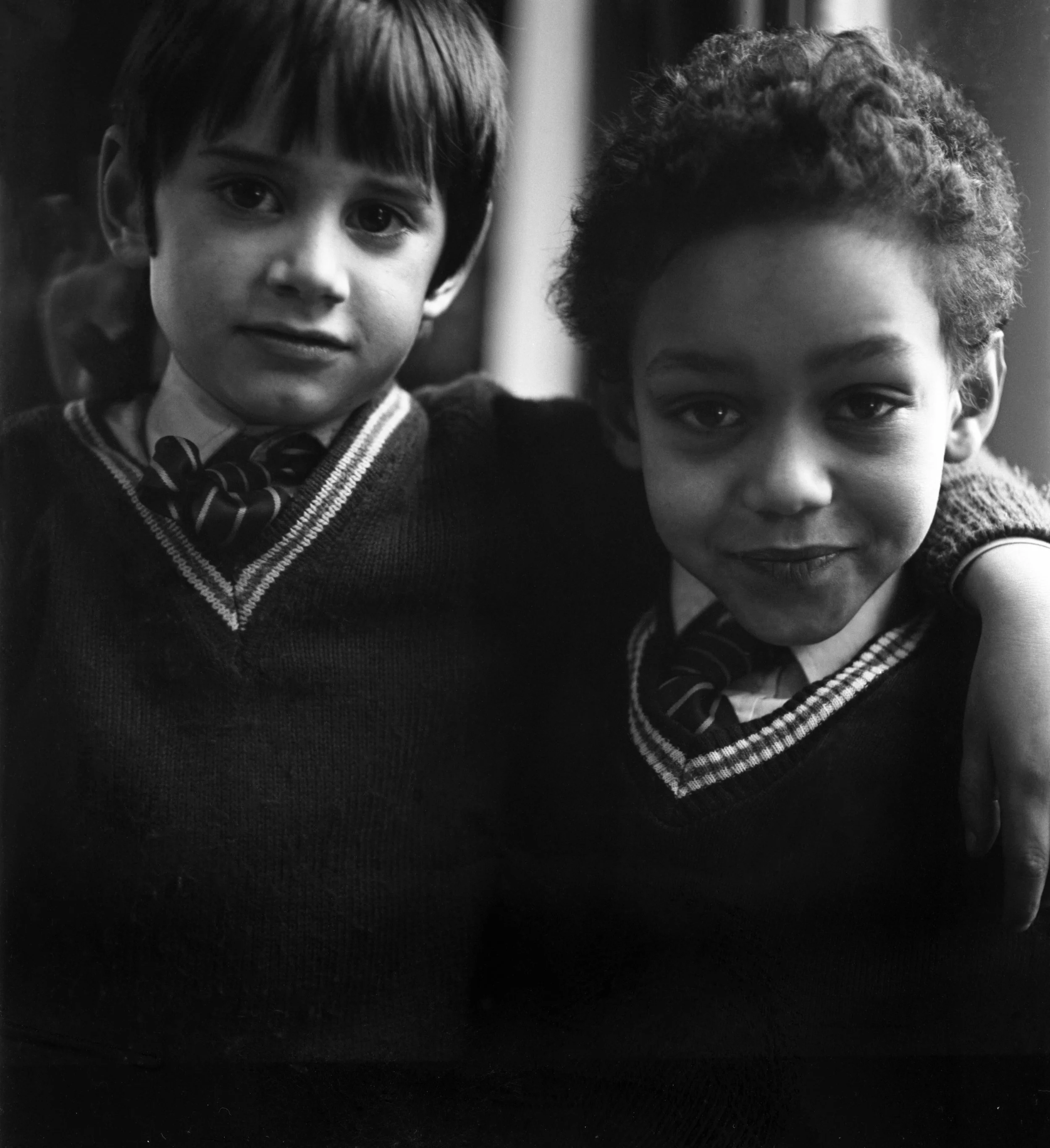 Black and white photo of two young boys. The boy on the left has straight dark hair and big eyes, wearing a collared shirt, tie, and sweater vest. The boy on the right has curly hair and a wide smile, wearing a sweater with a checkered collar. They a