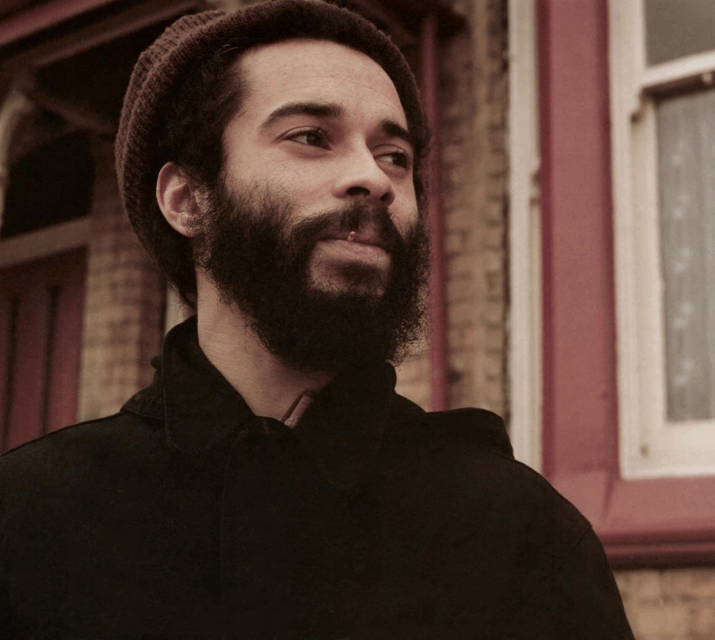 Close-up of a man with a full beard and thick eyebrows wearing a brown beanie and a black hoodie, standing outdoors near a building with brick walls and window frames.