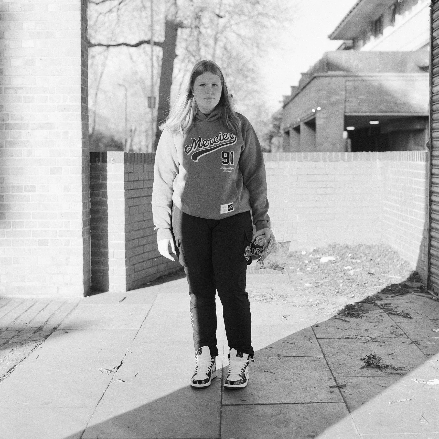 A woman wearing a 91 Mercury hoodie and athletic pants is standing outdoors on a sidewalk with ice skates in her hands. She is near a brick wall and a building, with trees and a fence in the background. The image is in black and white.
