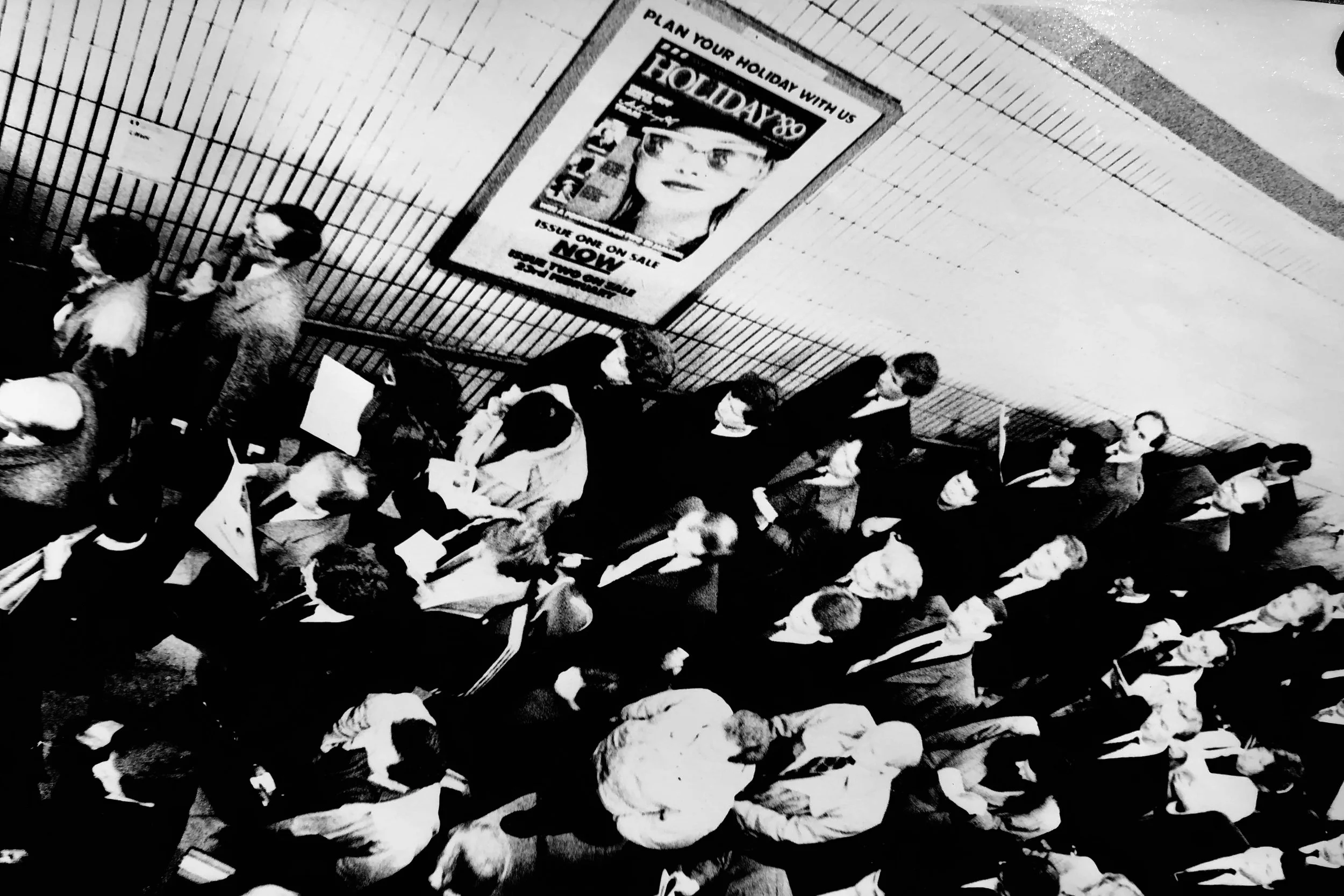 People lining up in front of a booth with a sign that reads "Plan Your Holiday with Us" and an advertisement for a magazine issue on sale.