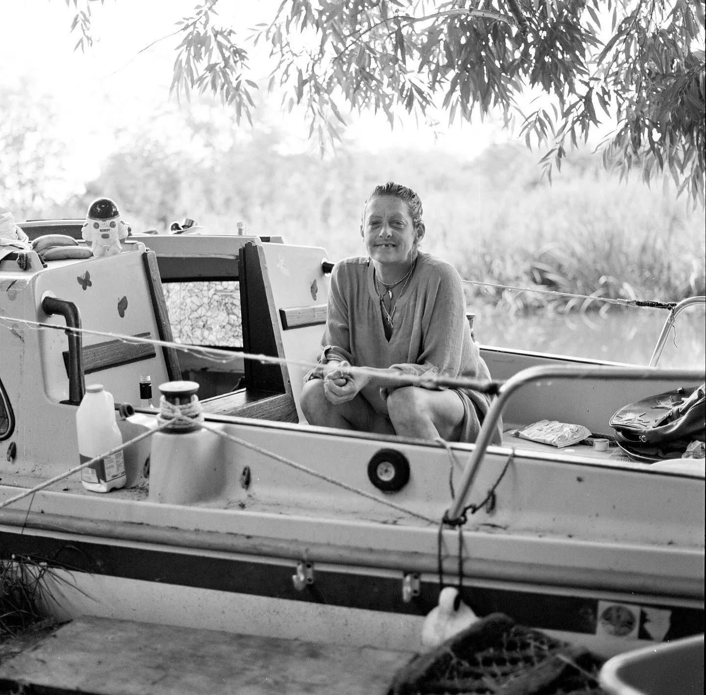 A woman sitting cross-legged in a small boat under trees, with various items on board, including a small toy astronaut and some bottles, in a black and white photo.