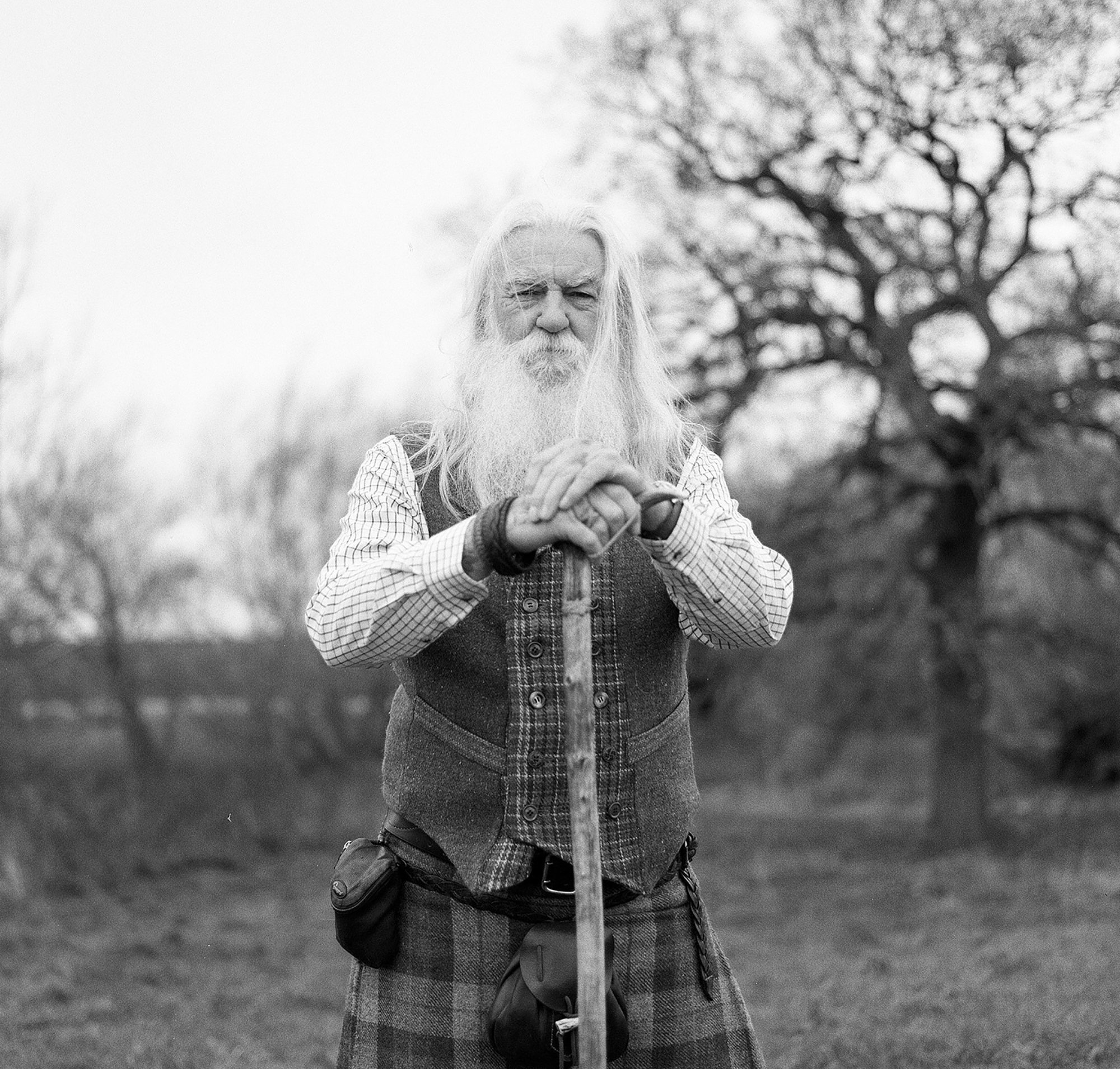 An elderly man with long white hair and a beard, wearing a plaid jacket, stands outdoors with a large tree in the background, holding a walking stick.