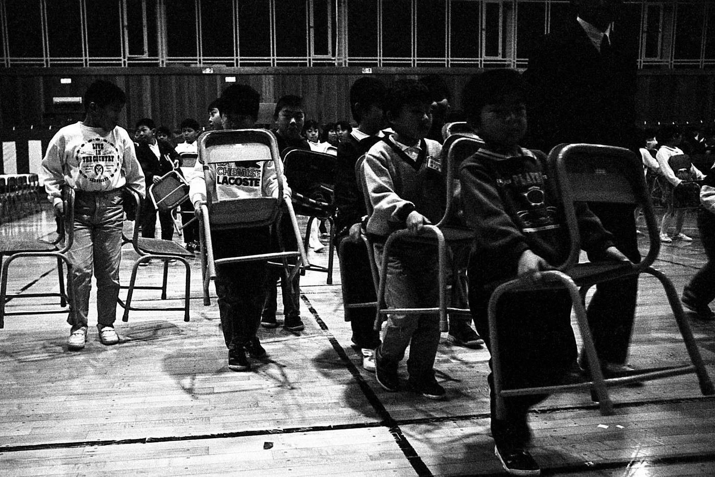 Children standing in line on a gymnasium floor, some holding chairs, in a school setting.