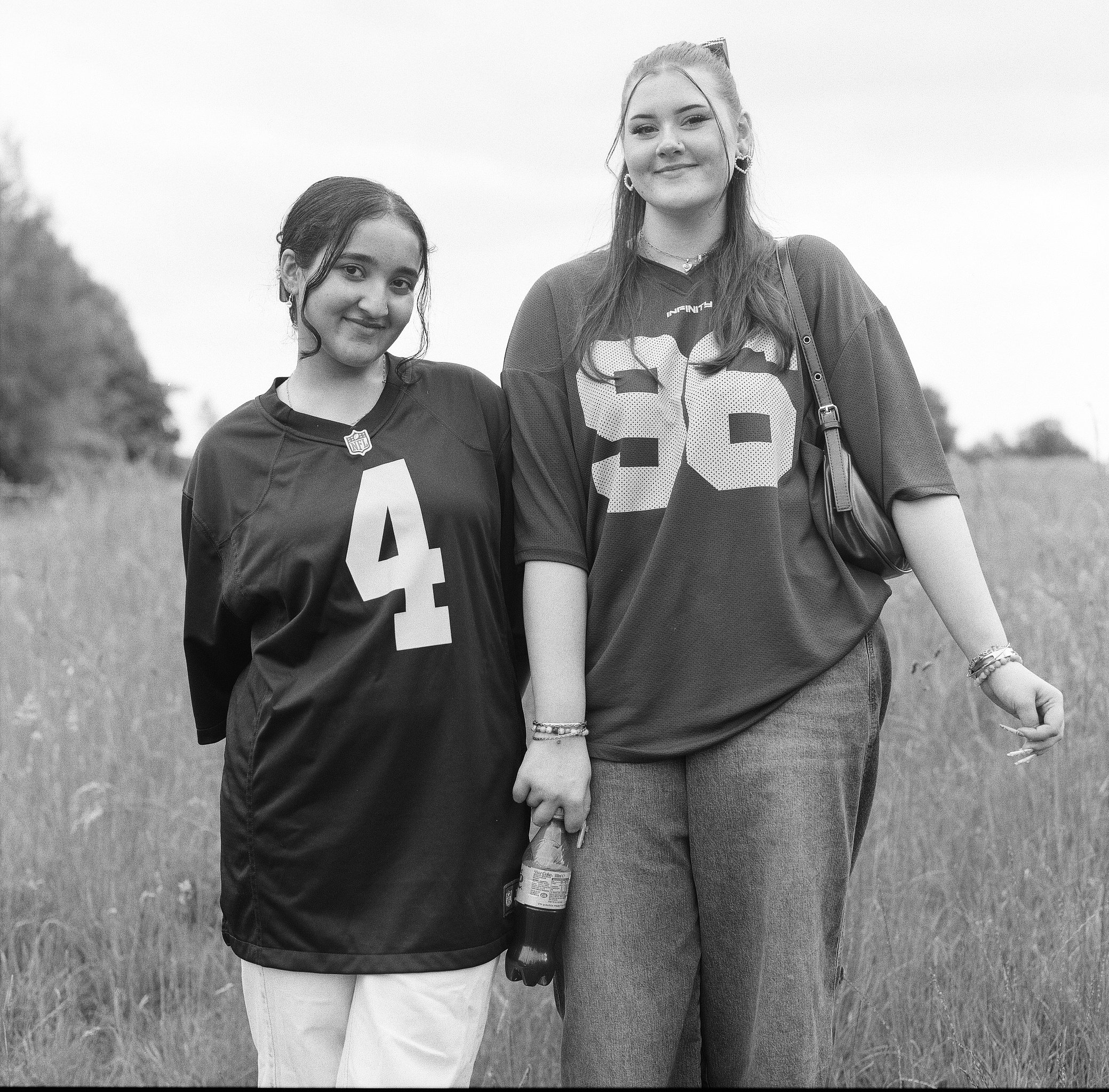 Two young women standing outdoors on a grassy field, smiling. One woman wears a sports jersey with the number 44 and an NFL logo, the other wears a patterned shirt and a cardigan, with various jewelry and a shoulder bag. The photo is in black and whi