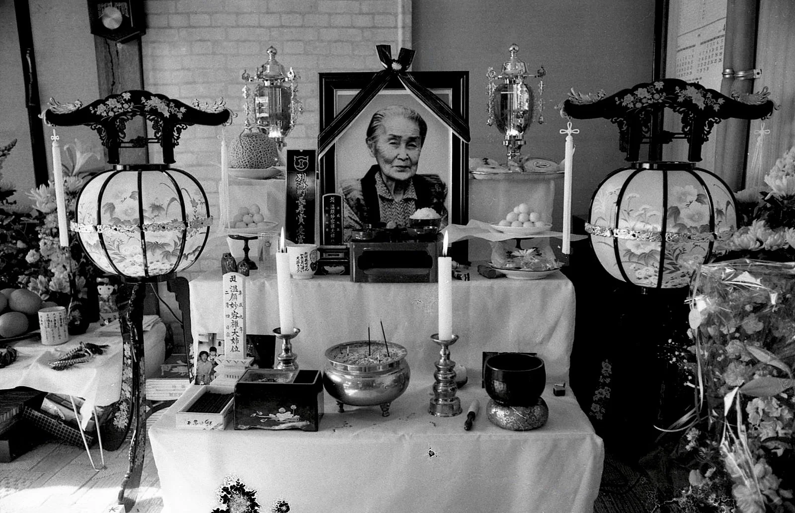 Black and white funeral altar with a framed photo of an elderly woman at the center. The altar has candles, incense, and offerings such as fruit and flowers. Traditional Japanese lanterns are positioned on either side of the altar.