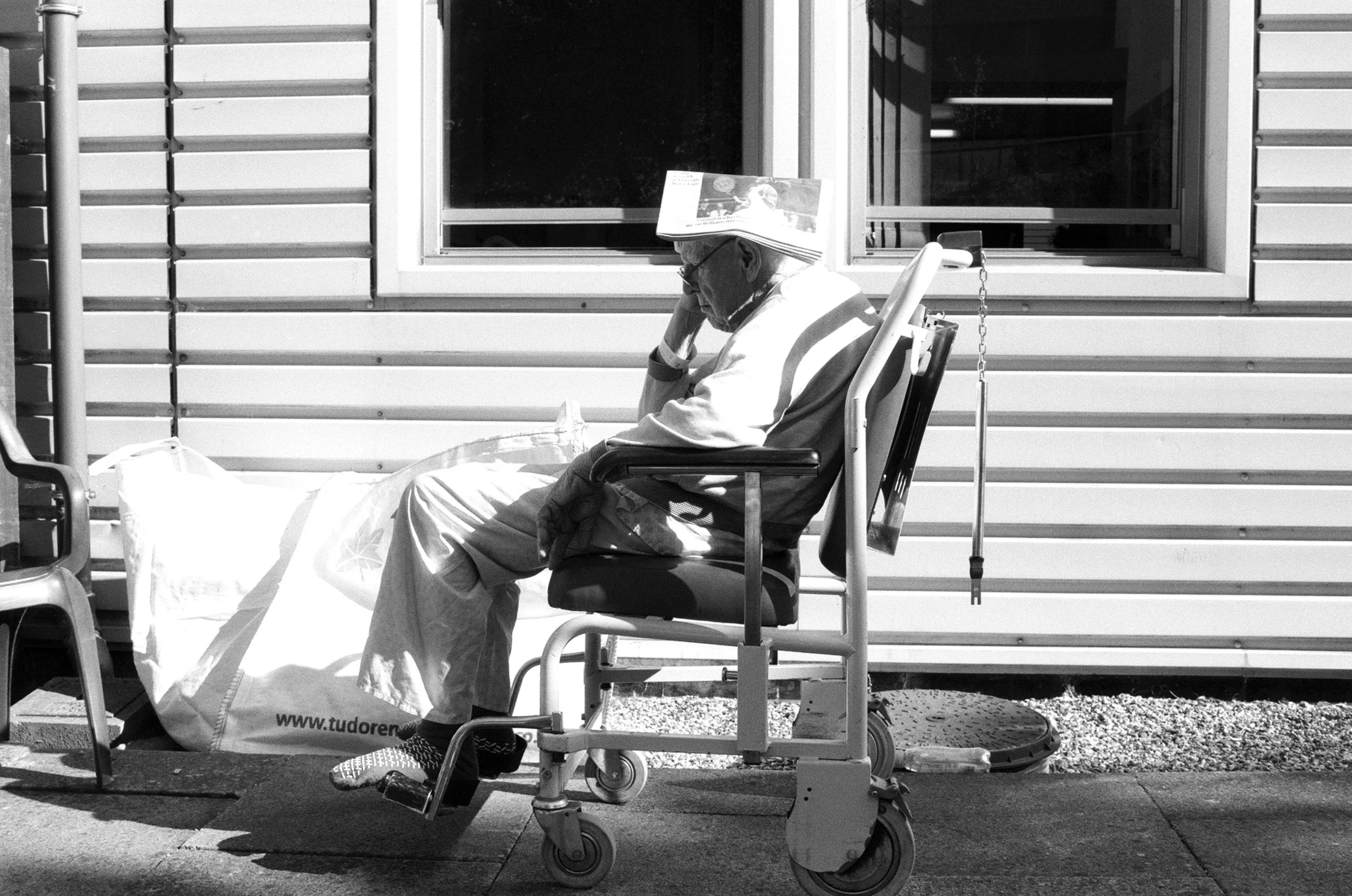 An elderly man wearing glasses and a hat reading a newspaper while sitting outside on a chair next to a wheelchair, in front of a building with horizontal siding.