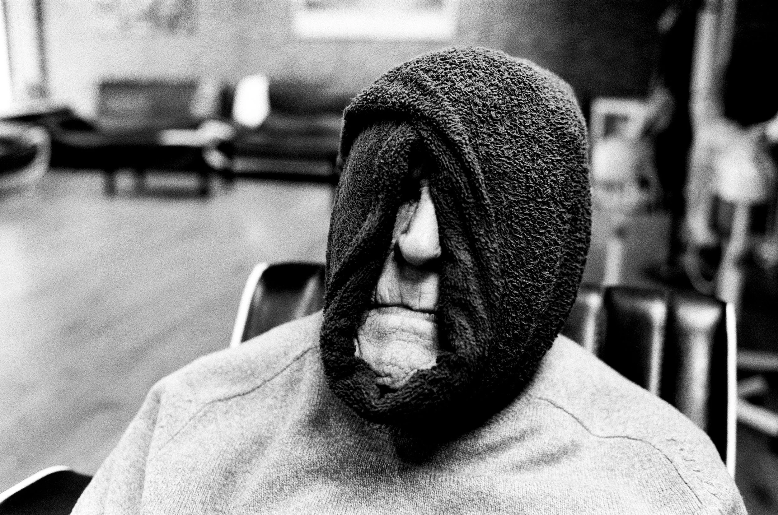A person with a towel wrapped around their head sits in a room with rows of chairs, wearing a sweater.