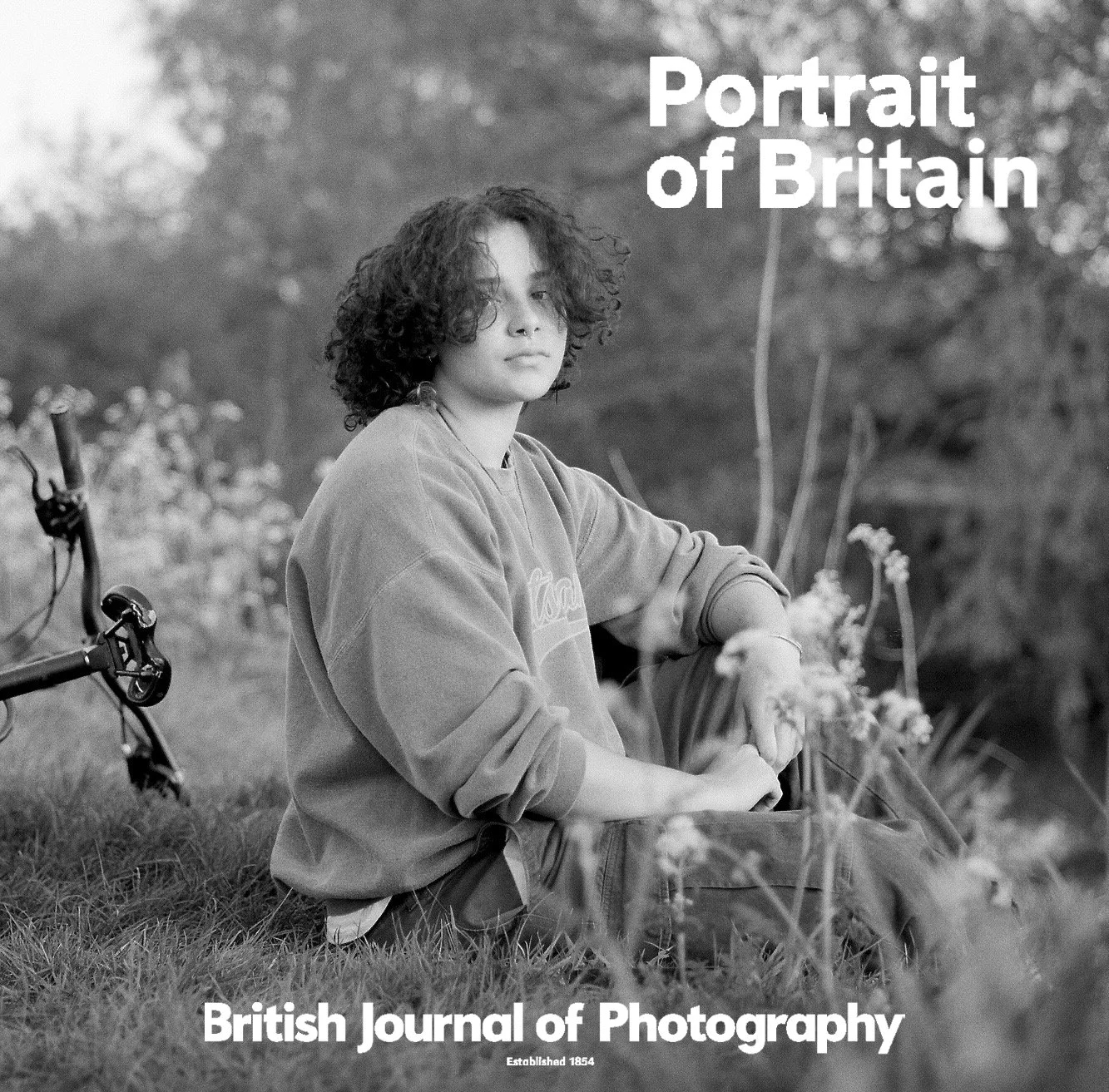 A black and white photograph of a young person with curly hair sitting outdoors in a natural setting, with a bicycle partially visible on the left side. The person is looking at the camera with a neutral expression, wearing a sweatshirt, and has a ba