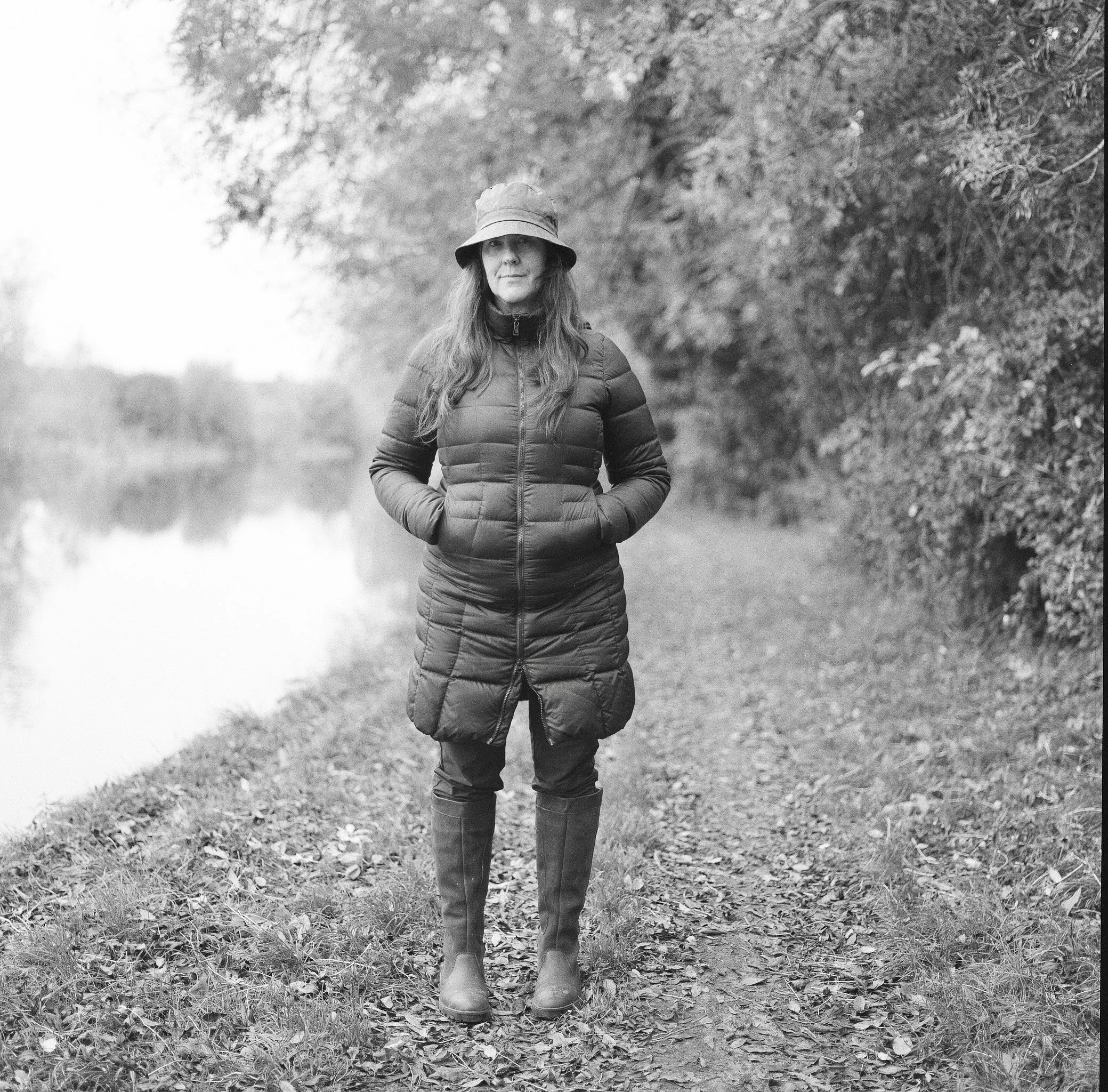 A woman in a puffy jacket, boots, and a wide-brim hat standing outdoors on a leaf-covered path surrounded by trees.