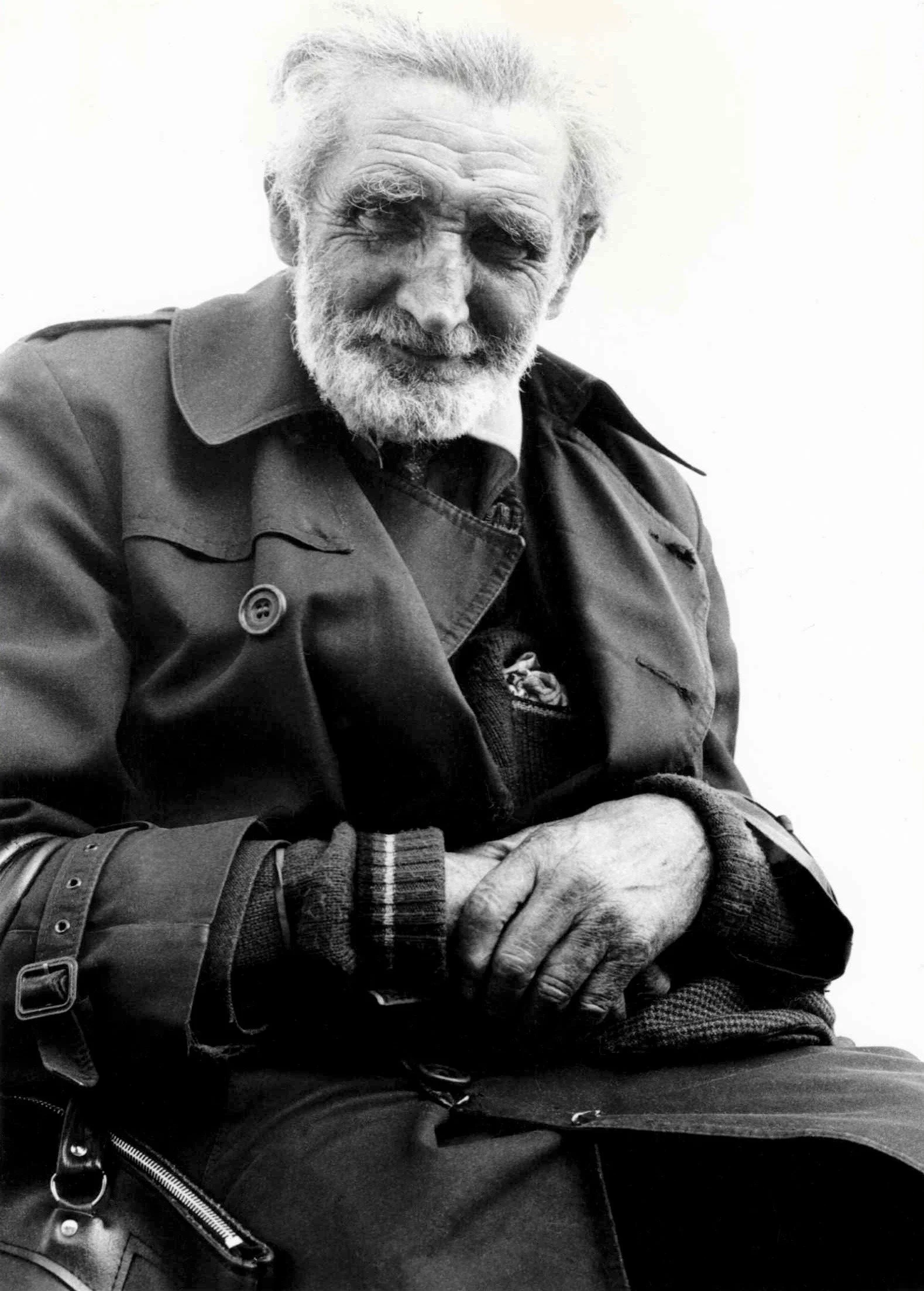 Black and white photo of an elderly man with a beard and mustache, wearing a jacket and a plaid shirt, sitting with his hands clasped in his lap.