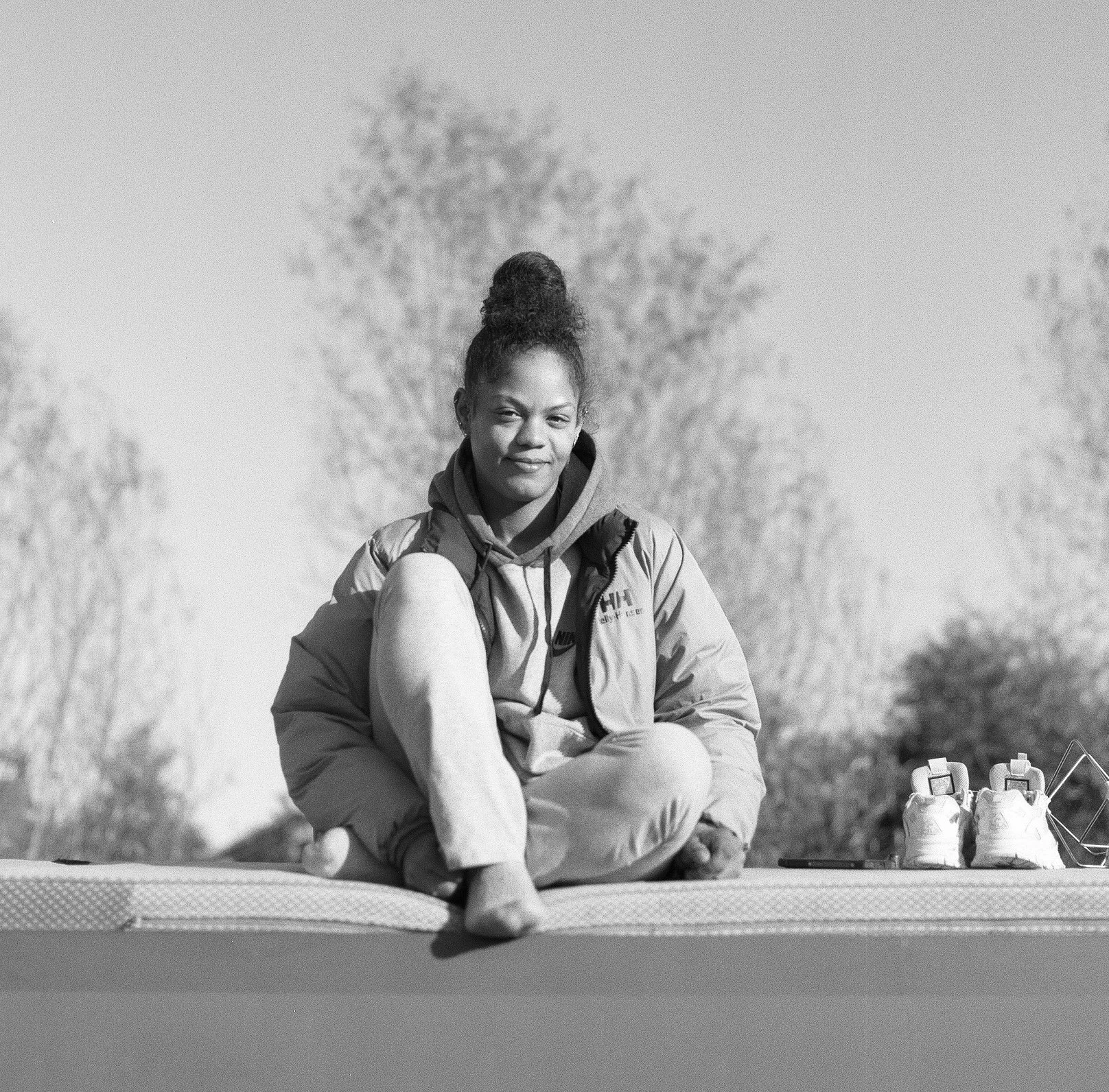 A woman sitting cross-legged outdoors on a yoga mat, with water bottles nearby, and trees in the background, in black and white.