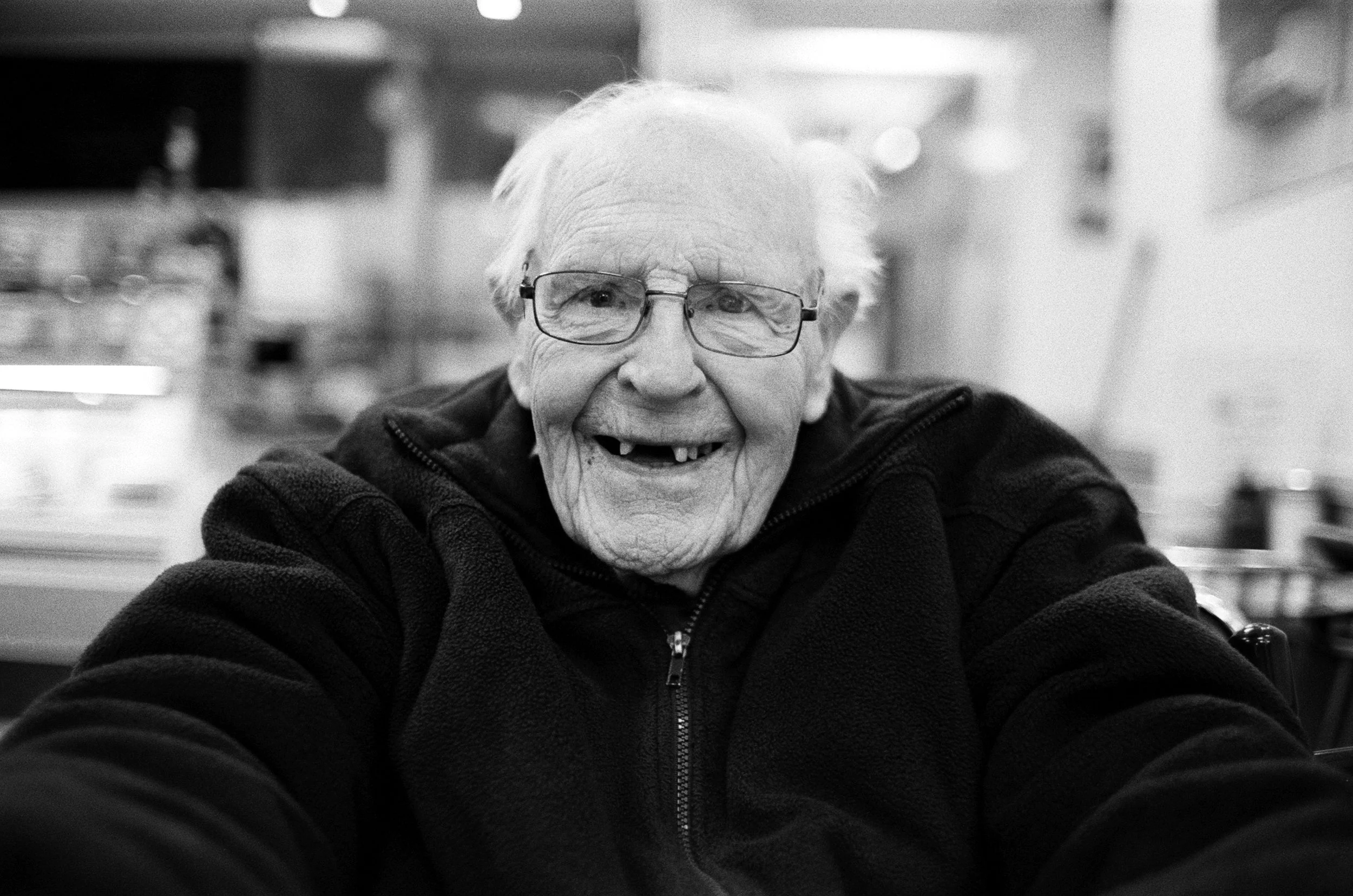 A smiling elderly woman with glasses, wearing a dark zip-up jacket, sitting indoors with a blurred background.