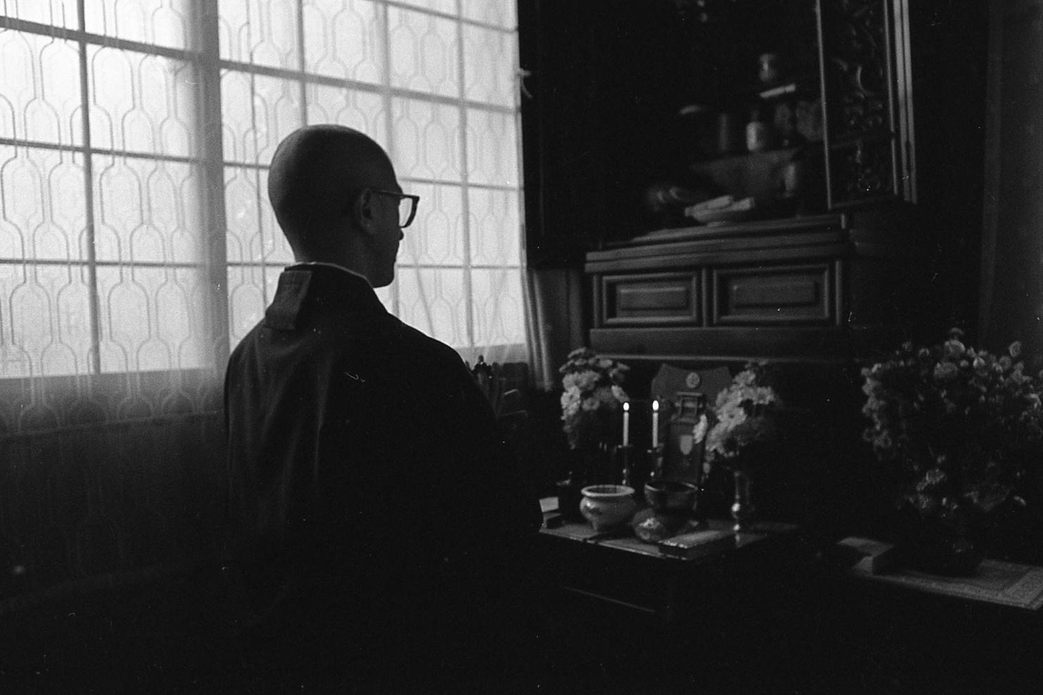 A black and white photograph of a man with glasses, sitting in front of an altar with candles, flowers, and a portrait, inside a room with lattice windows.
