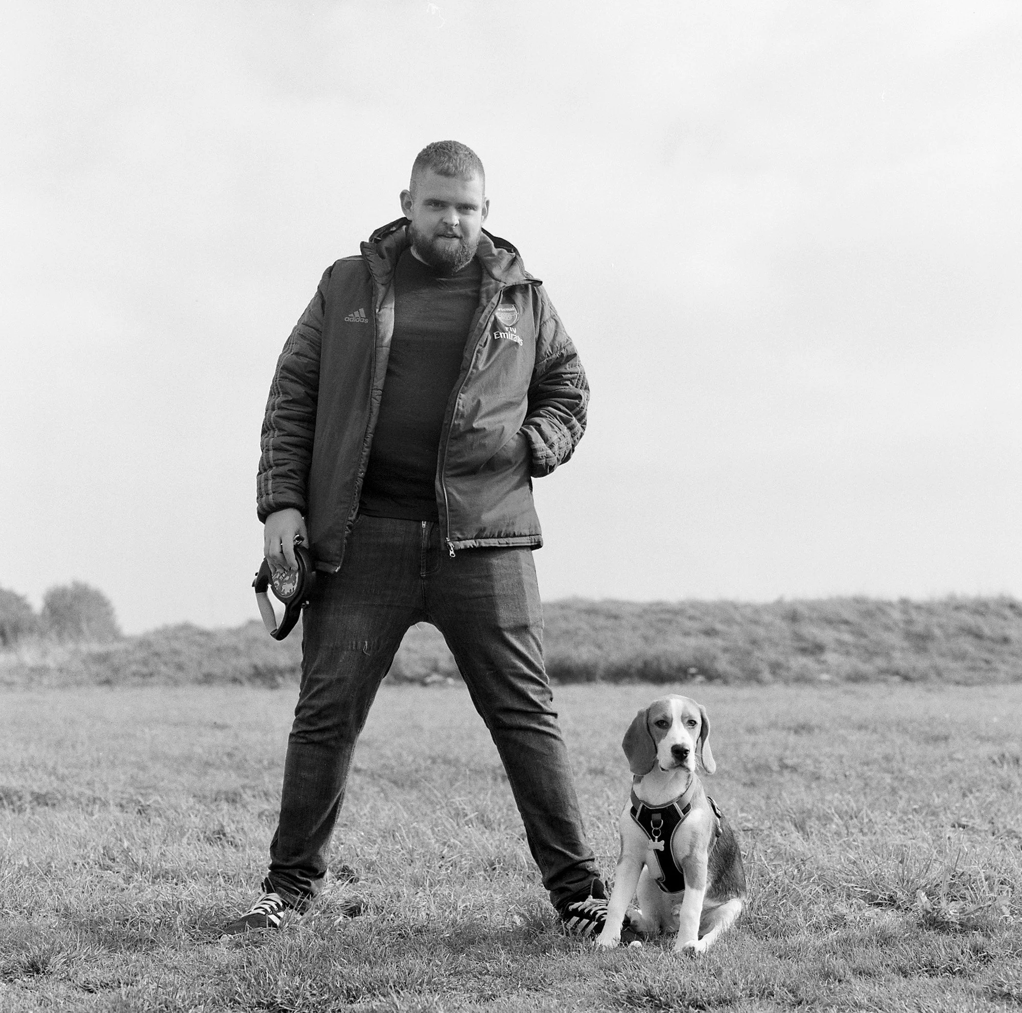 A man with a beard wearing a dark T-shirt and a puffy jacket stands outdoors in a grassy field, holding a leash, with a beagle dog sitting on the ground beside him. The man is casually dressed and looking at the camera.