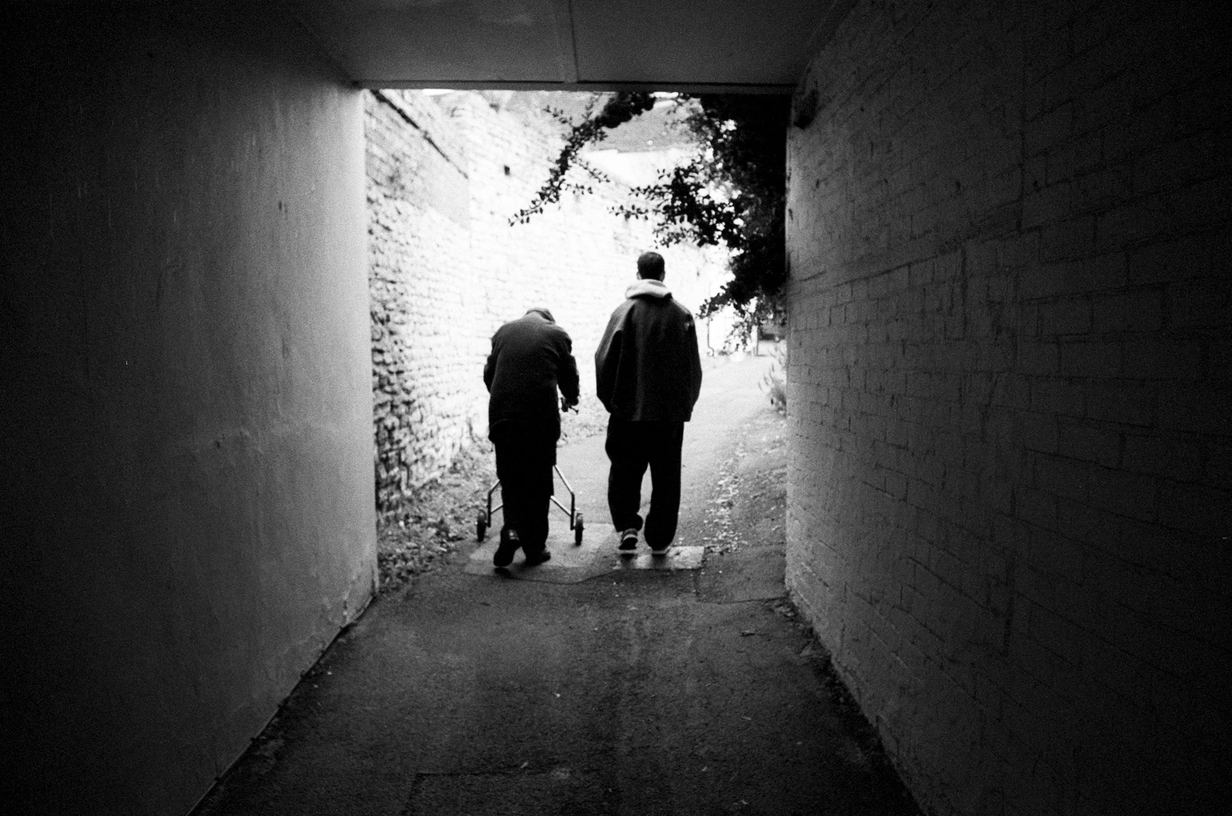 Two people, one of whom is using a walker, walk through a dark underpass toward a brighter outdoor area with trees and a building in the background.