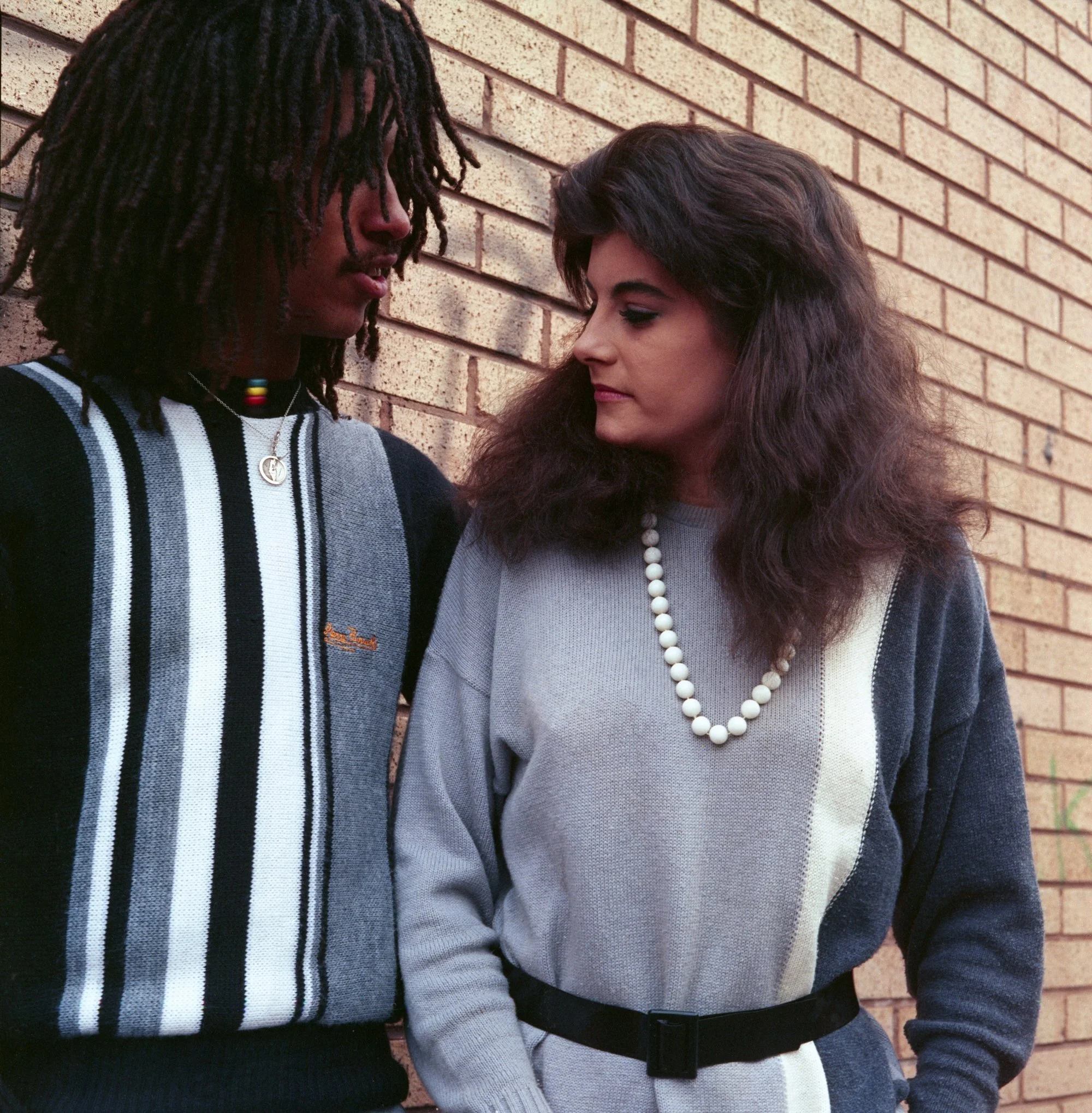 Close-up of two people standing close to each other against a brick wall, one with dreadlocks and the other with wavy hair, both with serious expressions.