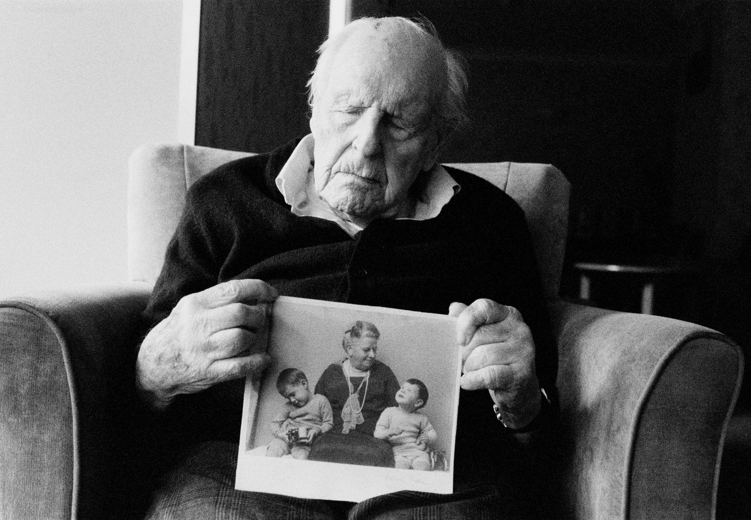An elderly man sitting in a chair reading a book that features a black-and-white photo of children.