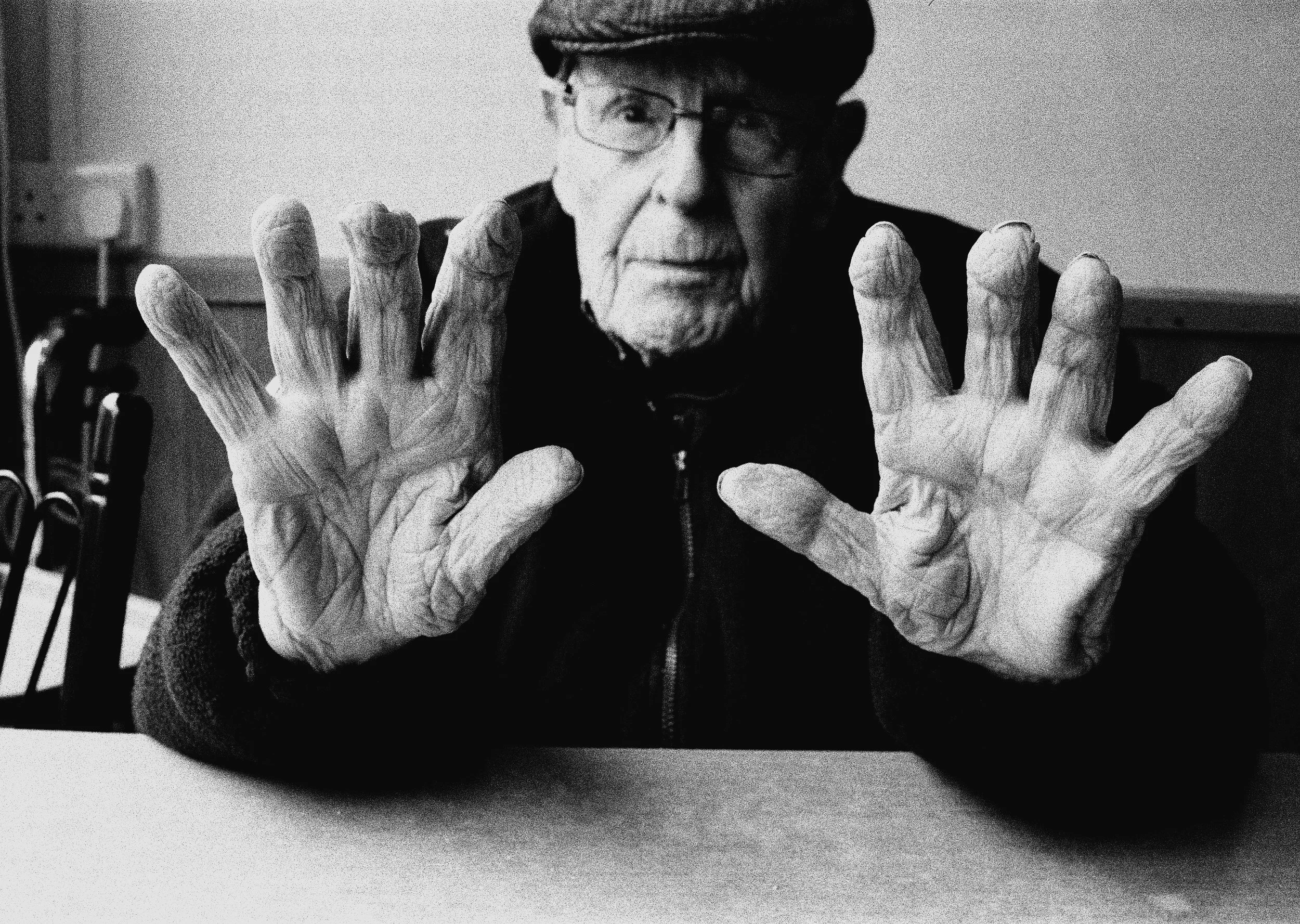 Black and white photo of an elderly person with glasses, wearing a cap, raising their hands towards the camera, with a blurred background.