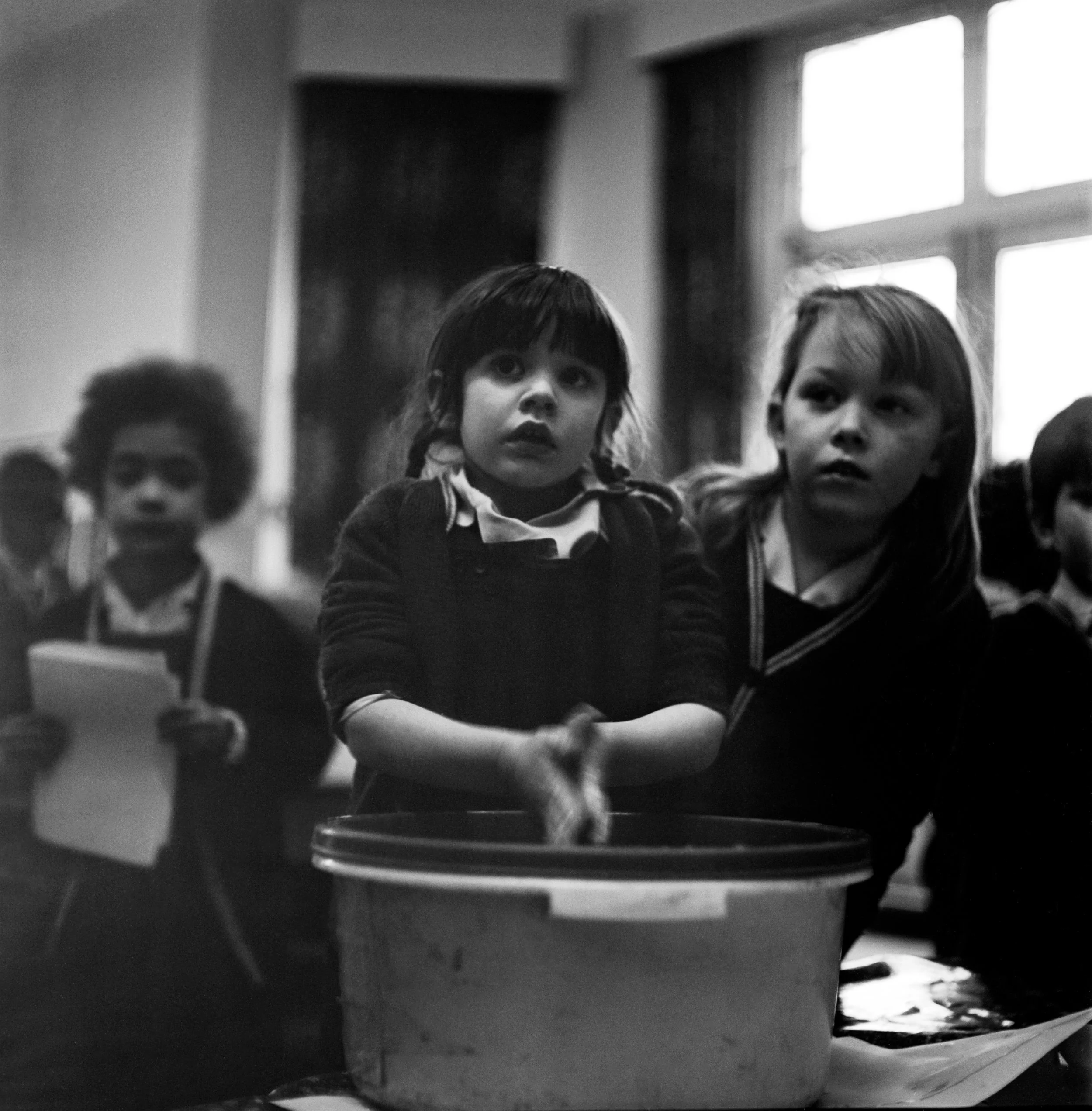 Black and white photo of children sitting in a classroom, with one girl in the center looking upward, and others nearby, surrounded by desks and windows.