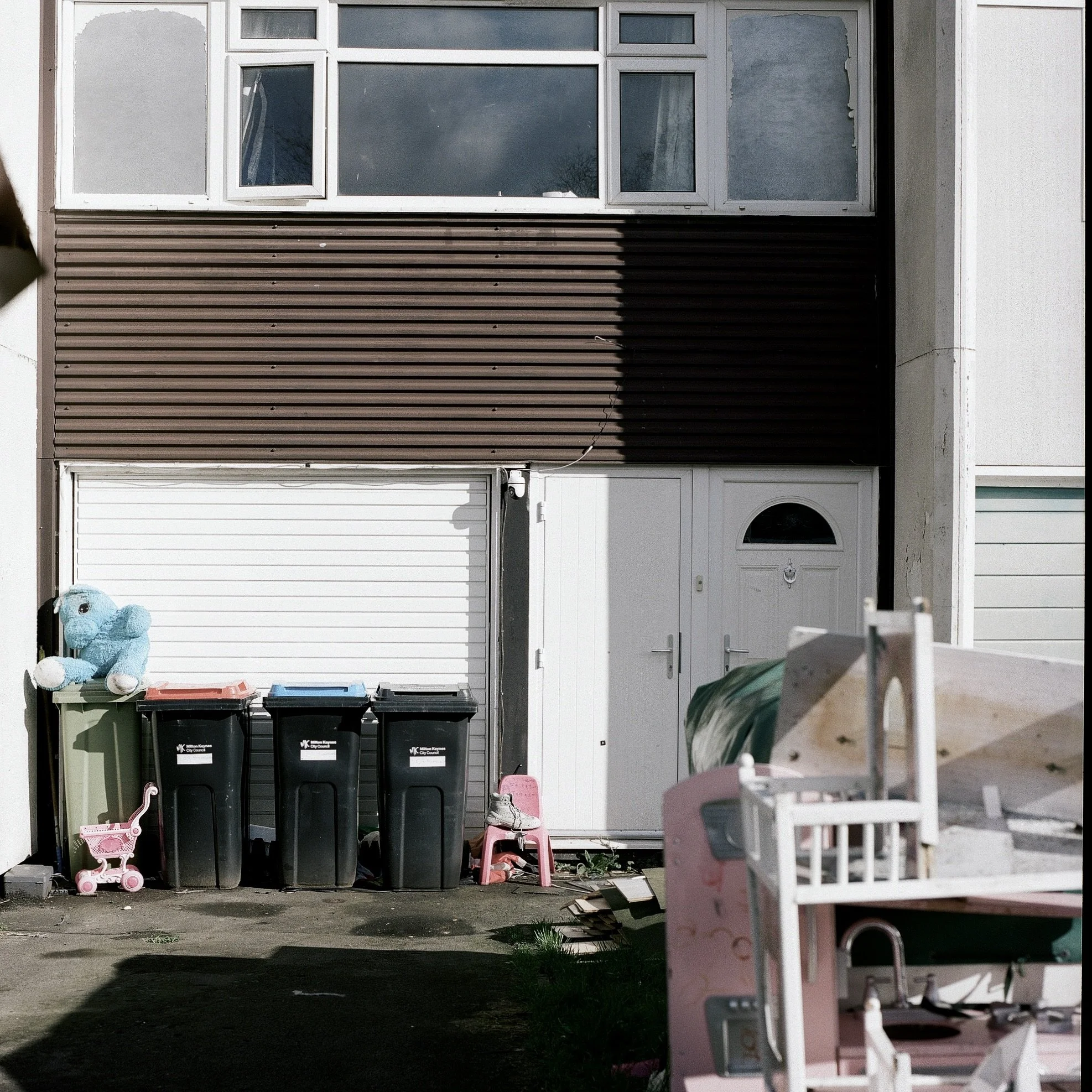 A residential house with a closed garage door, a white front door, and windows. In front of the house, there are three black trash bins, a pink doll-sized stroller, a pair of baby shoes, and a pink toy shopping cart. Shadows and sunlight are visible 