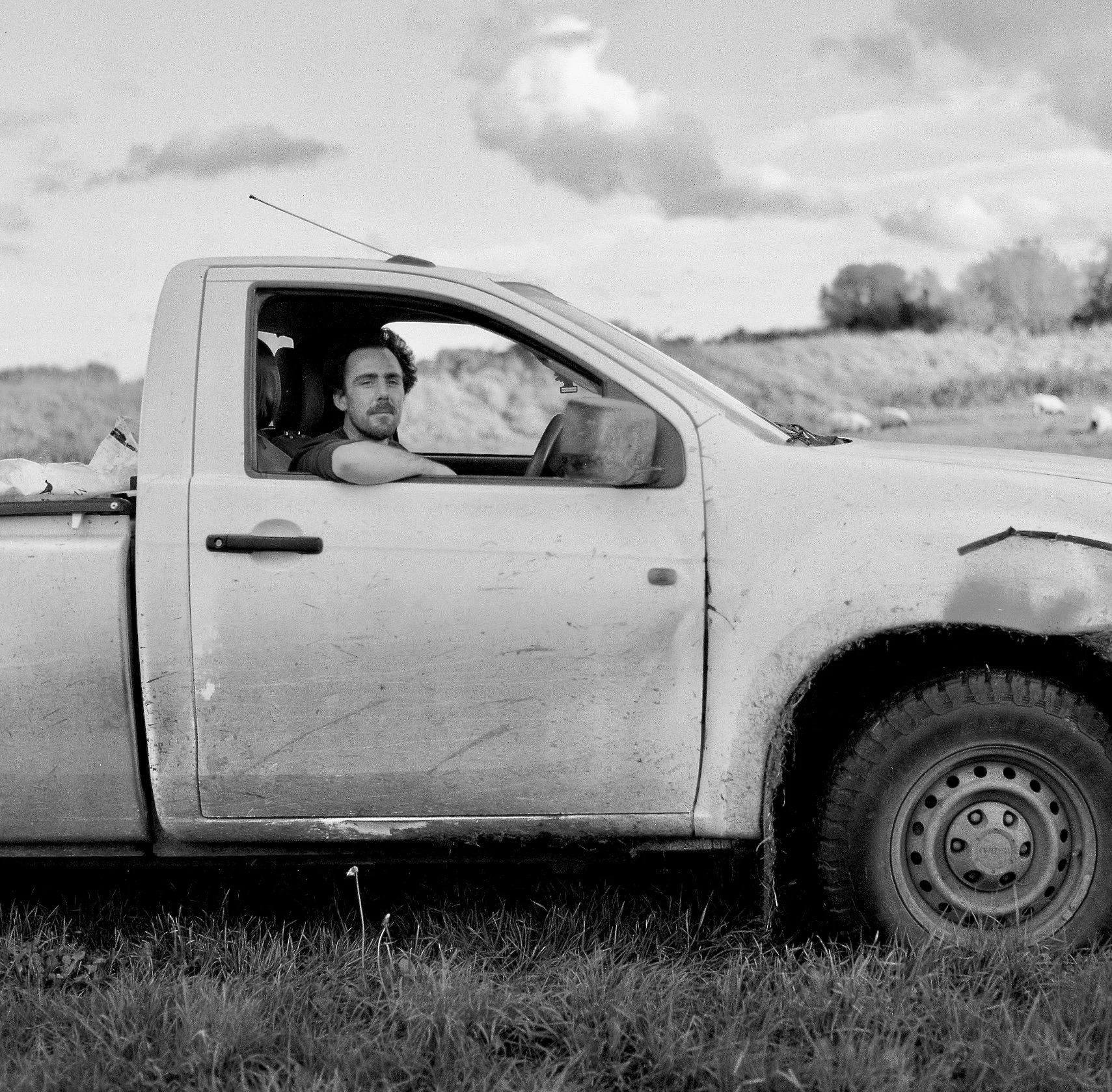 Black and white photo of a man with dark hair and beard sitting in the driver's seat of a pickup truck, looking out the window. The truck is parked on grass near a vast open landscape with fields, distant trees, and a cloudy sky.