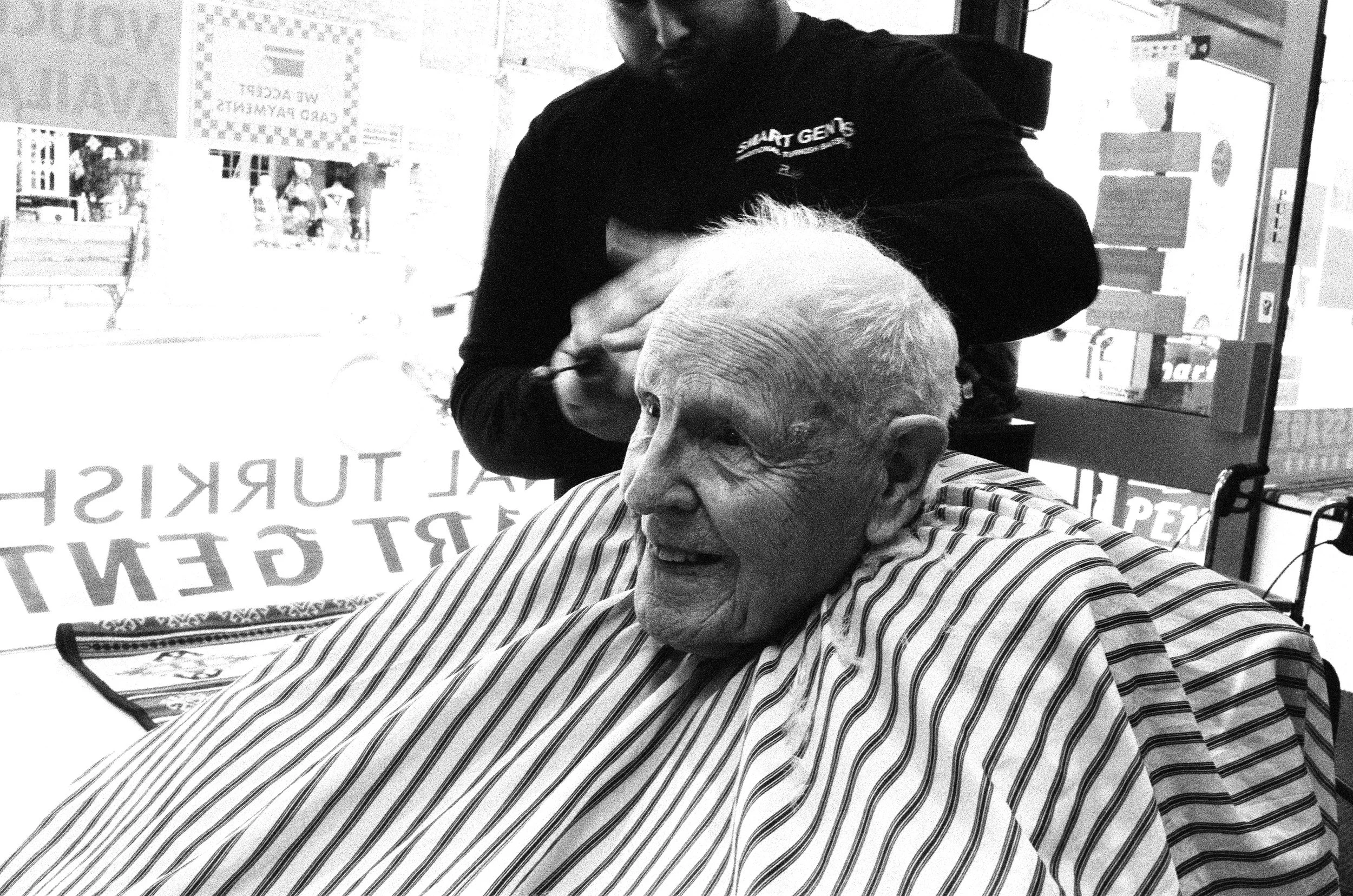 Older man with light-colored hair and striped shirt sitting at a barber shop chair receiving a haircut from a bearded barber. The barber is wearing a dark shirt with a logo and is focused on cutting the man's hair. The scene is in a barbershop with a