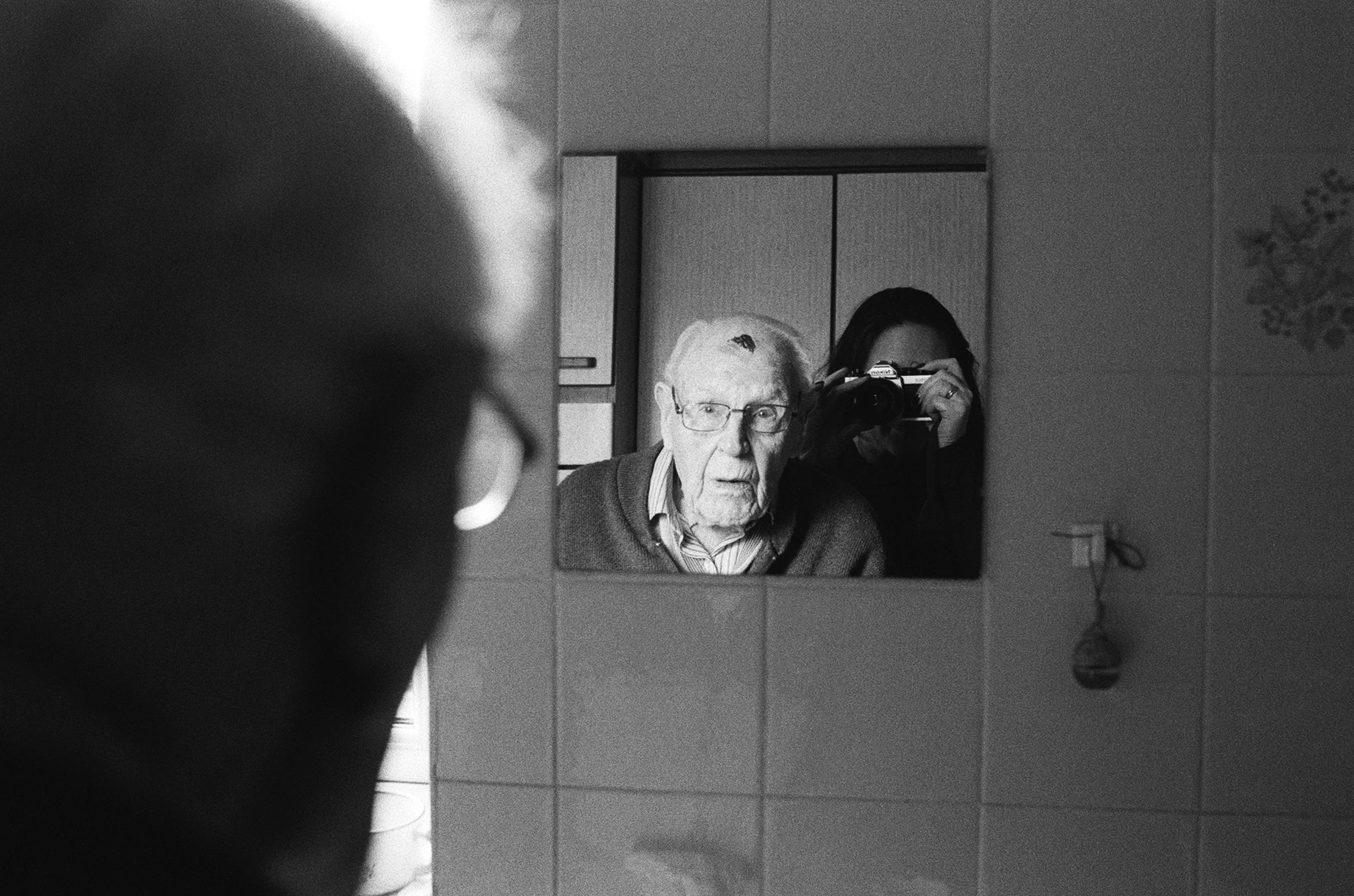 An elderly man with glasses and a striped shirt looking into a mirror, with a woman holding a camera behind him taking the photo. The setting appears to be a bathroom or a room with tiled walls and a decorative wall sticker of a plant.