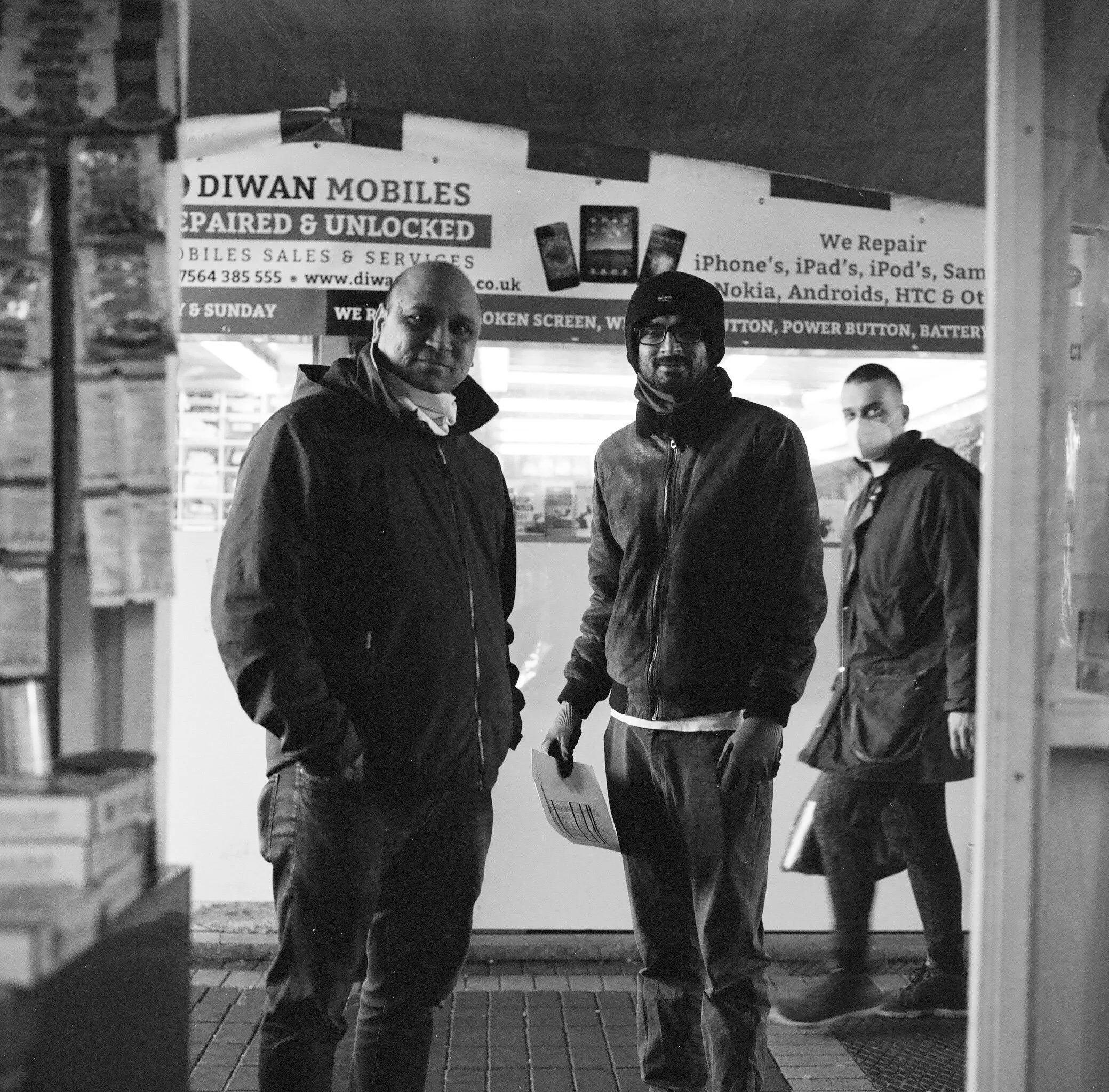 Two men standing outdoors in front of a sign for a phone repair shop. One man is bald and wearing a dark jacket, while the other has dark hair, glasses, and a beanie. Other people can be seen in the background.
