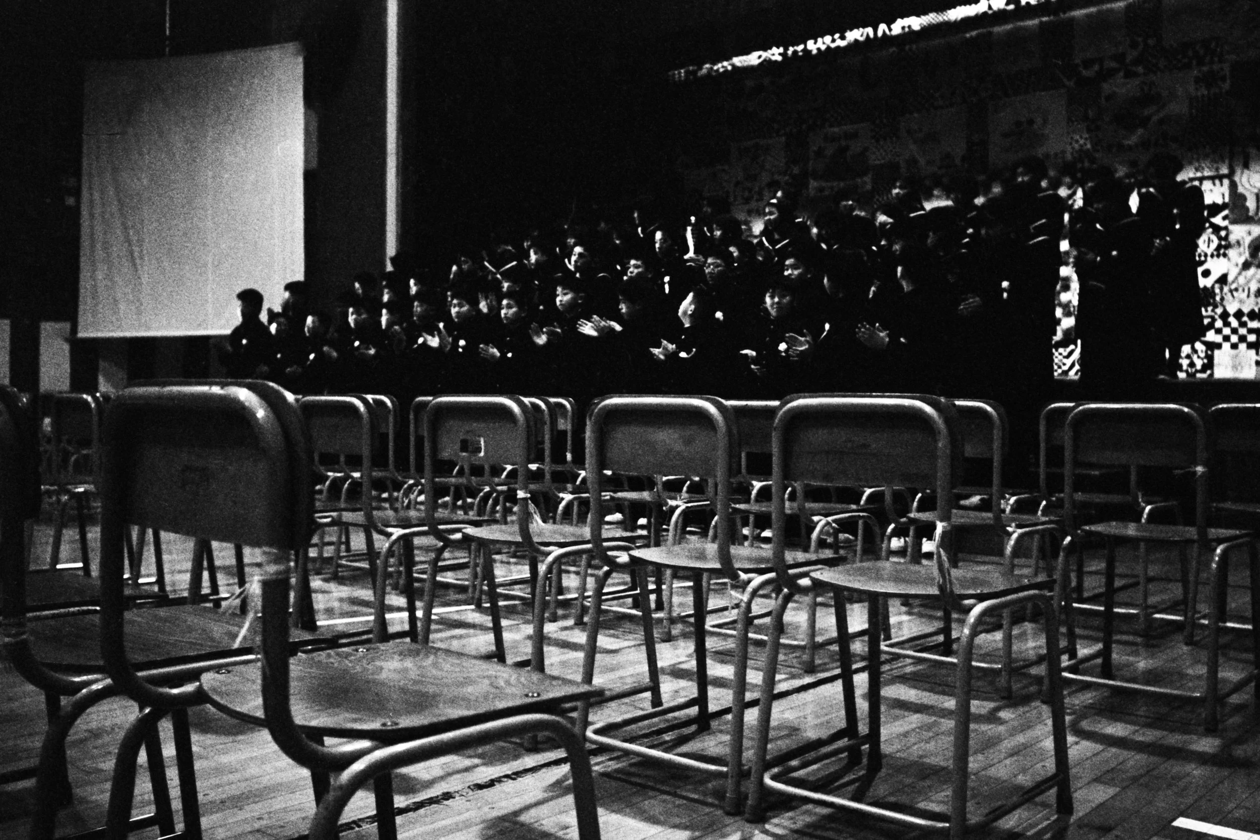 A black and white photo of an auditorium or theater with empty chairs and a group of children or students sitting on a stage, clapping and facing the audience.