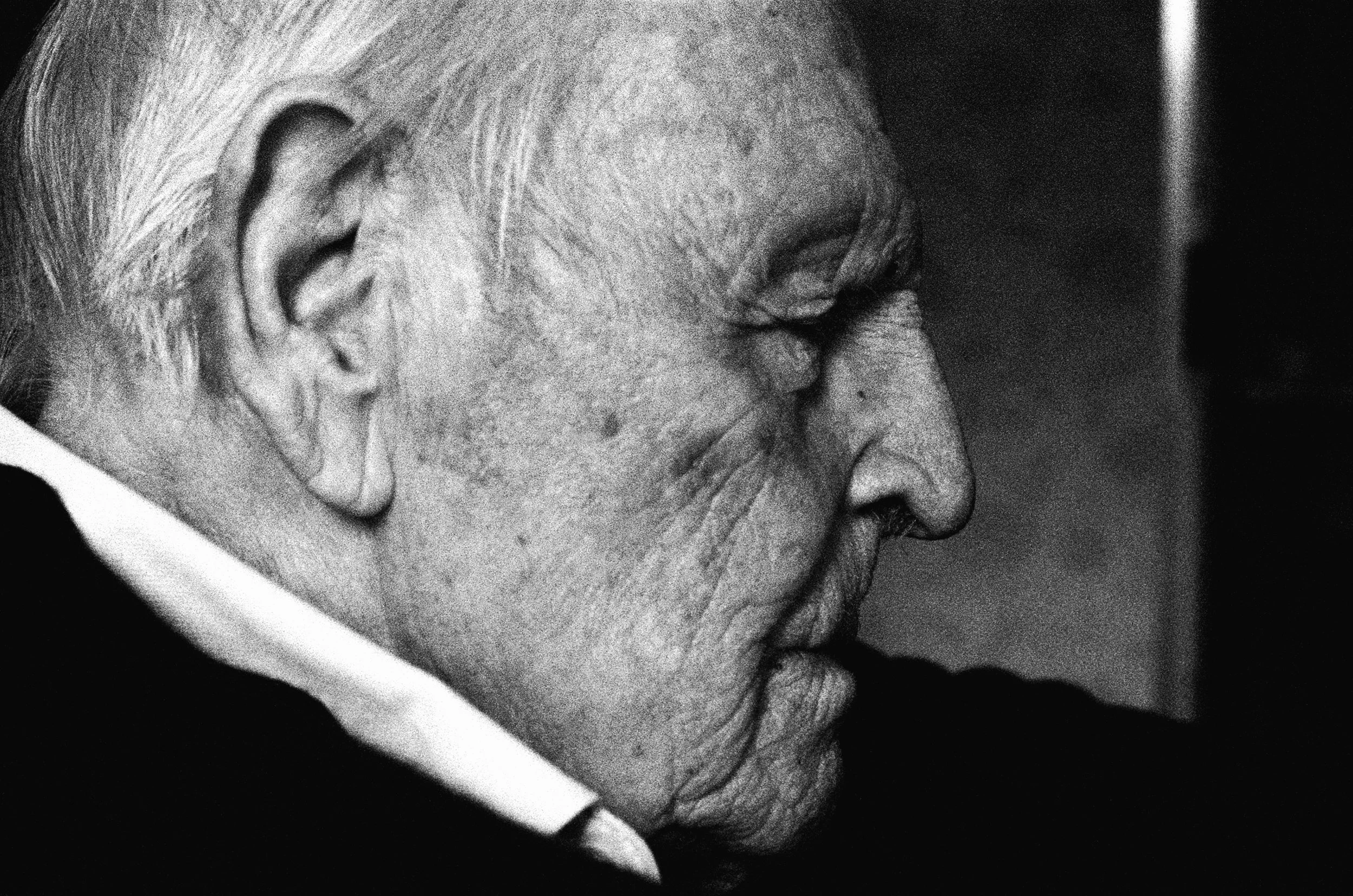 A close-up black and white photograph of an elderly man with white hair, wearing a suit with a white collar, looking down with his hand resting against his face.