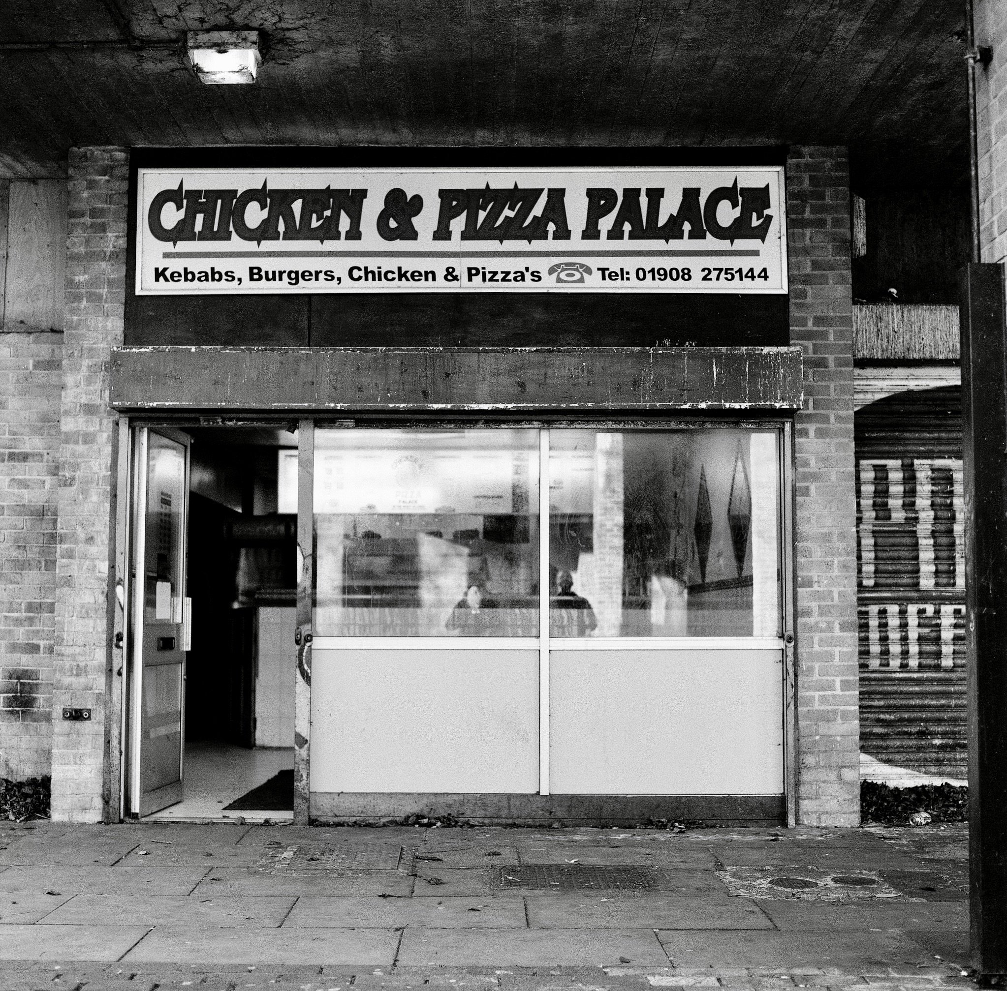 Black and white photo of a building entrance with a large sign reading 'Chicken & Pizza Palace.' The sign also lists menu items including kebabs, burgers, chicken, and pizza, and displays a telephone number. The entrance has an open door with glass p