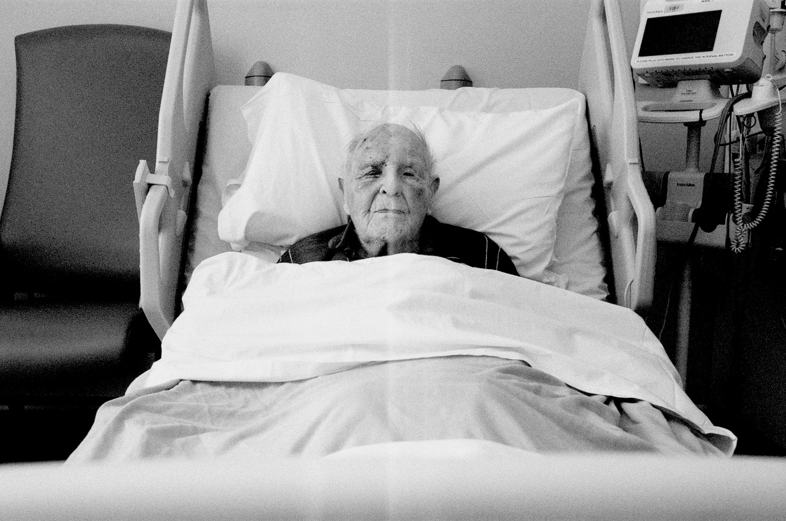 An elderly man with a shaved head and closed eyes lying in a hospital bed with medical equipment nearby.