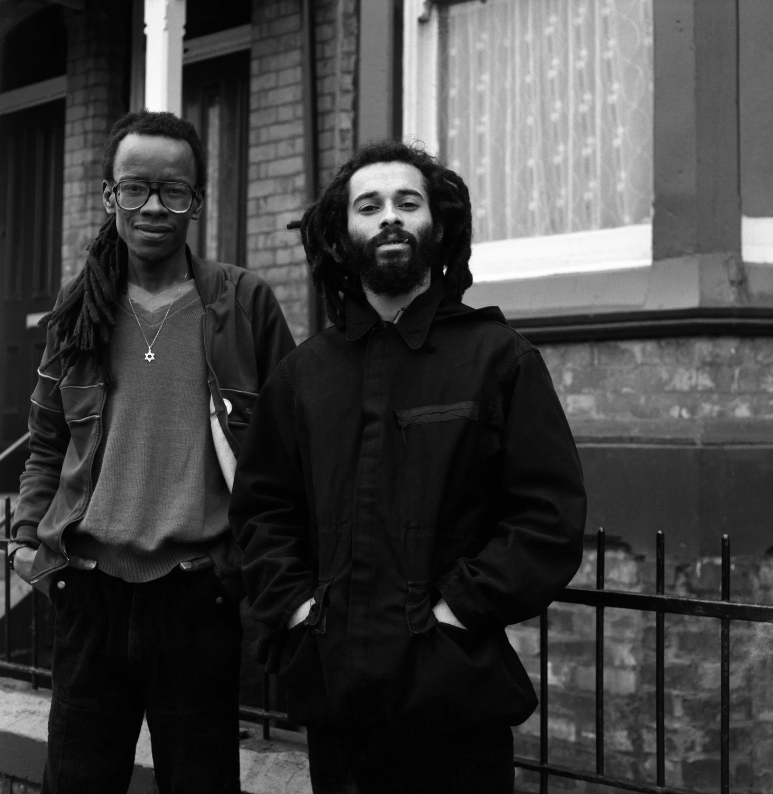 Two men with dreadlocks and beard standing outdoors in front of a brick building and metal fence. One man wears glasses and a star necklace, the other wears a dark jacket. Black and white photograph.