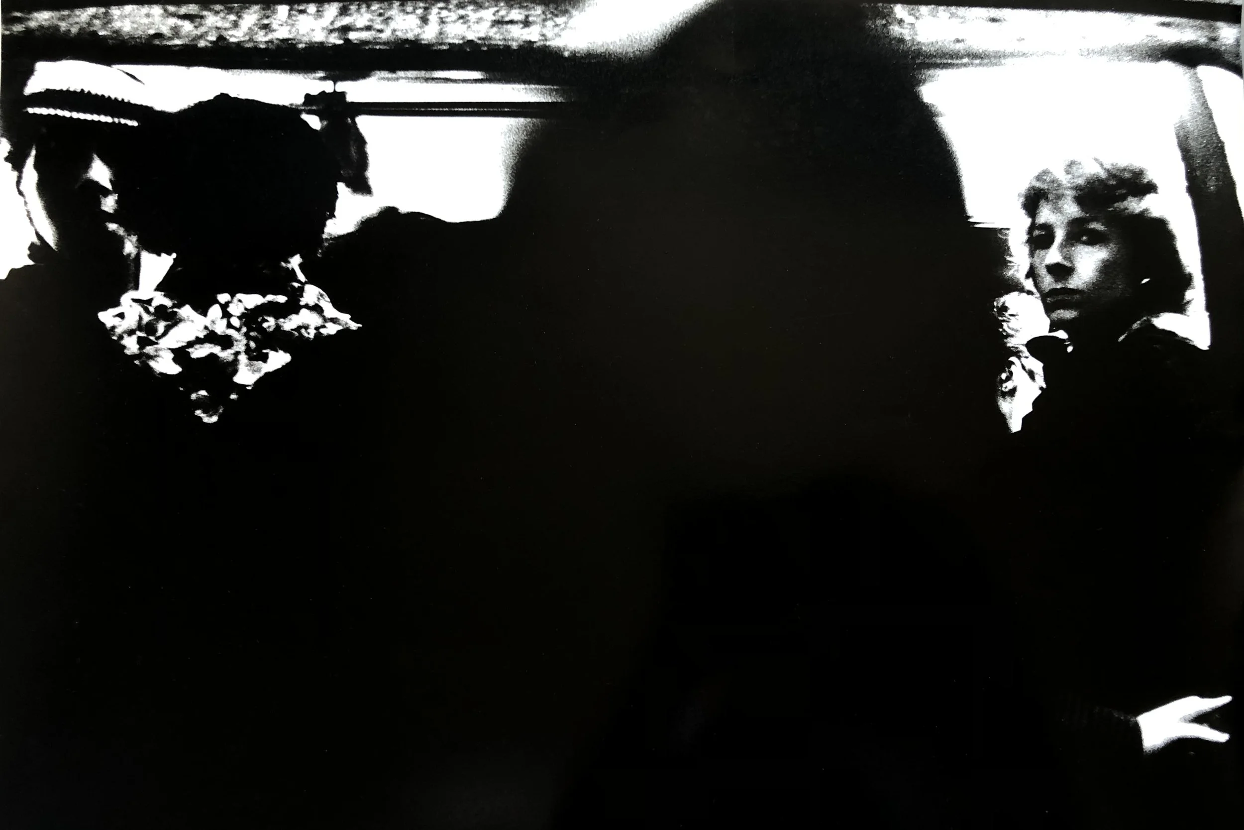 Black and white high-contrast photograph of two women, with one on the left wearing a floral dress and a hat, and the other on the right with curly hair and a serious expression, indoors near a window.
