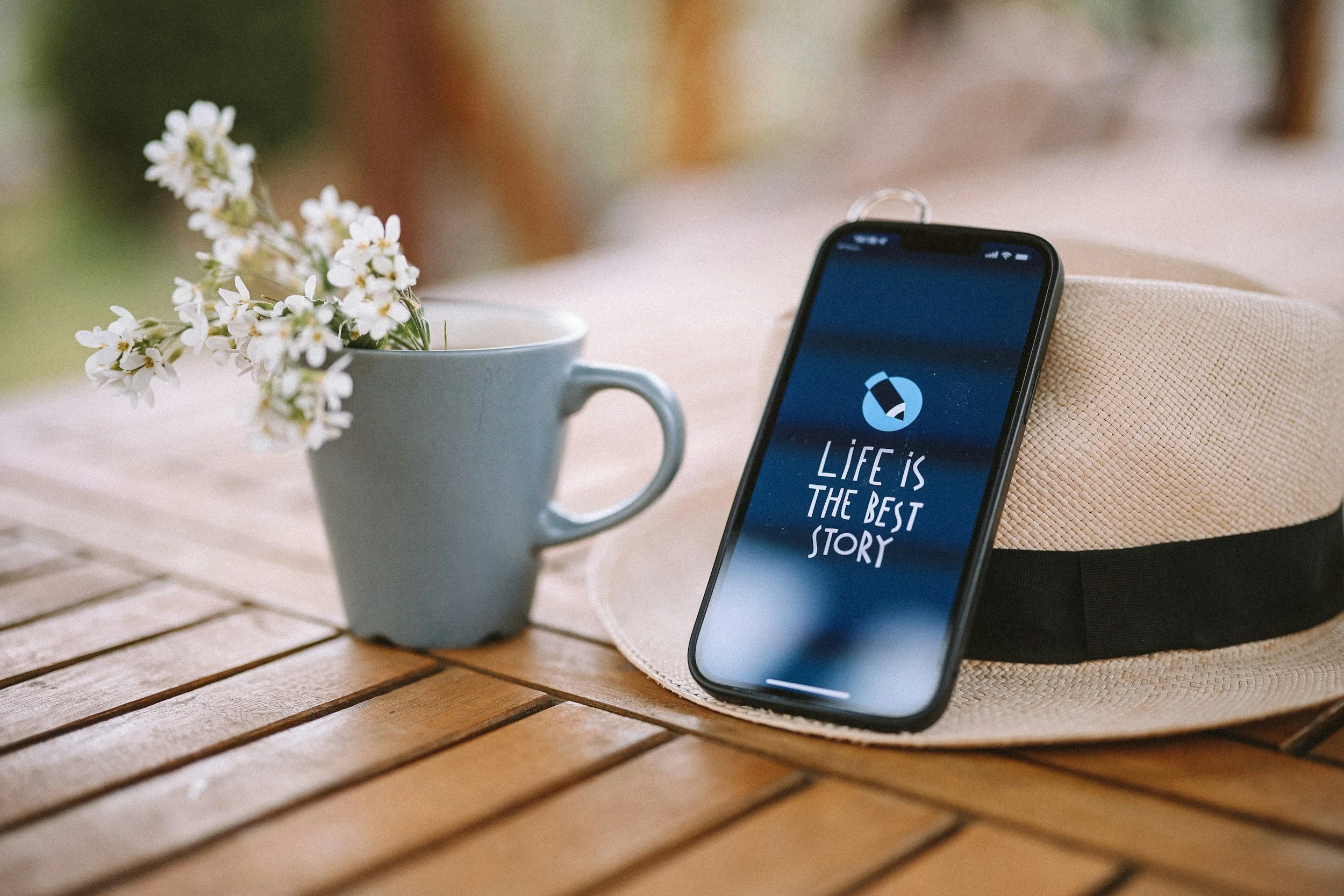 A table with a light blue mug filled with white flowers, a smartphone displaying a quote, and a straw hat with a black ribbon.