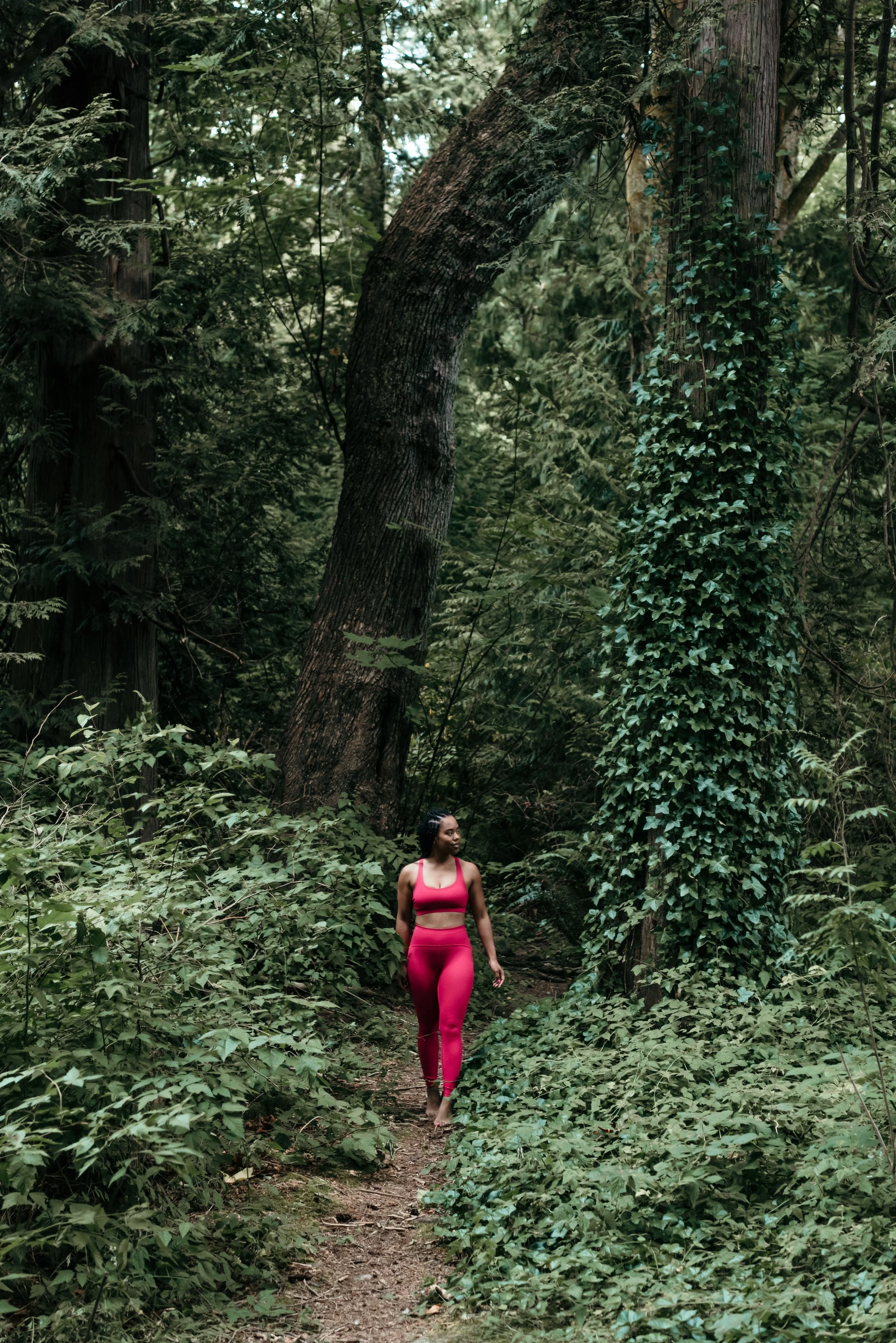 A woman in pink athletic wear walking on a narrow trail in a dense, green forest filled with tall trees and thick foliage.