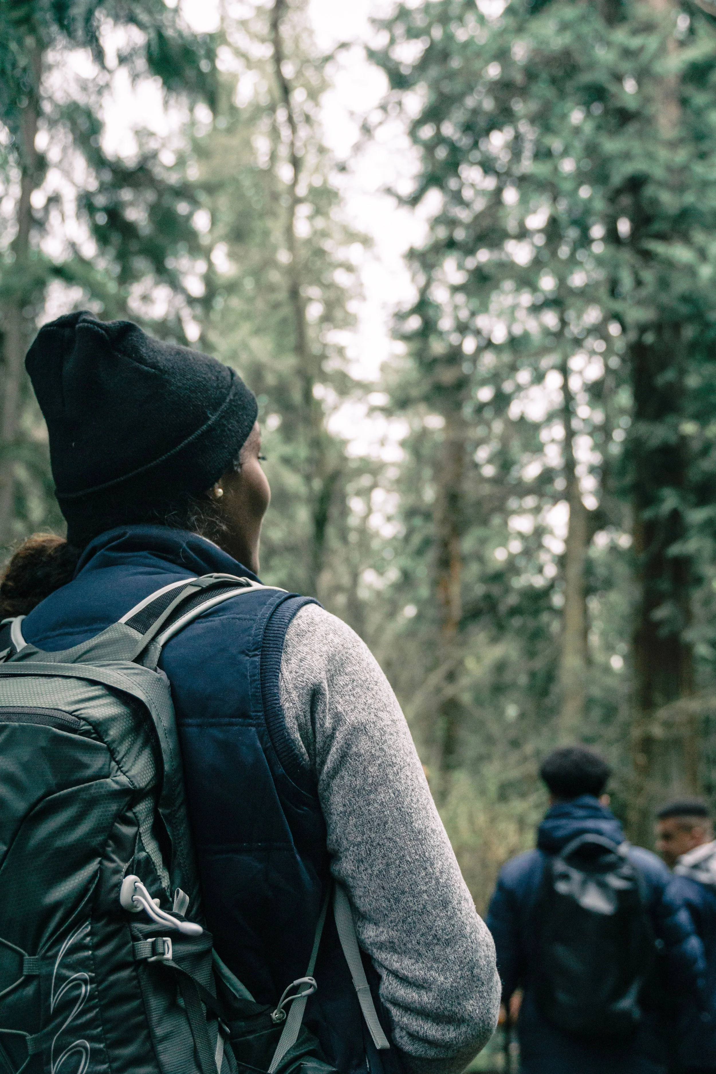 A woman with a black beanie and a backpack, and two men with backpacks, are standing outdoors in a forested area with tall trees and green foliage.