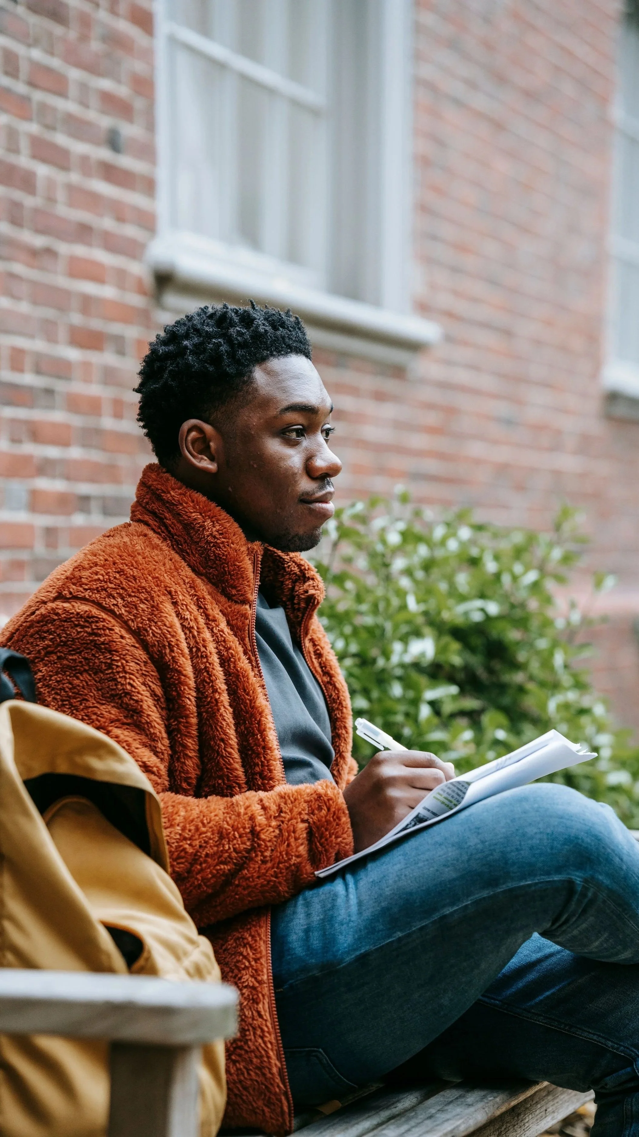 Young man with dark curly hair sitting on a bench outdoors, writing in a notebook, wearing an orange fuzzy jacket and blue jeans, with a brick building and green bushes in the background.