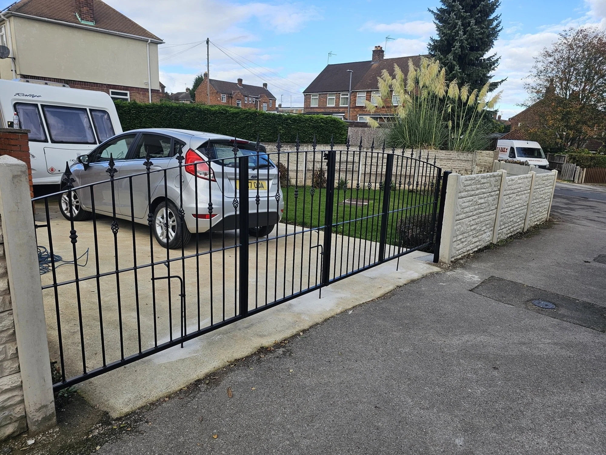 A residential driveway with a black metal gate, gray car, and a white caravan, surrounded by a stone wall and a garden with tall grasses and trees, under a partly cloudy sky.