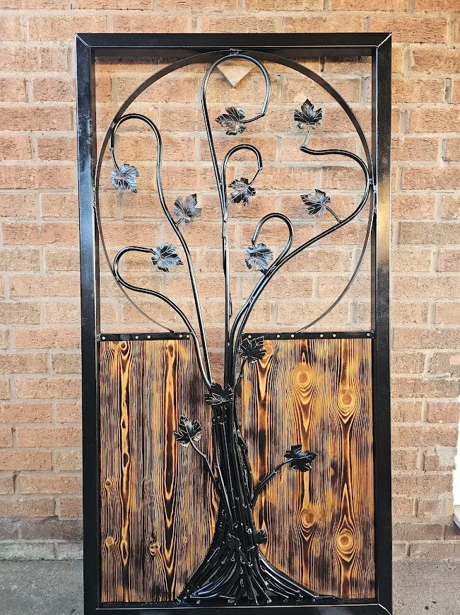 Decorative metal tree sculpture with leaves, mounted on a brown wood and black metal frame against a brick wall.